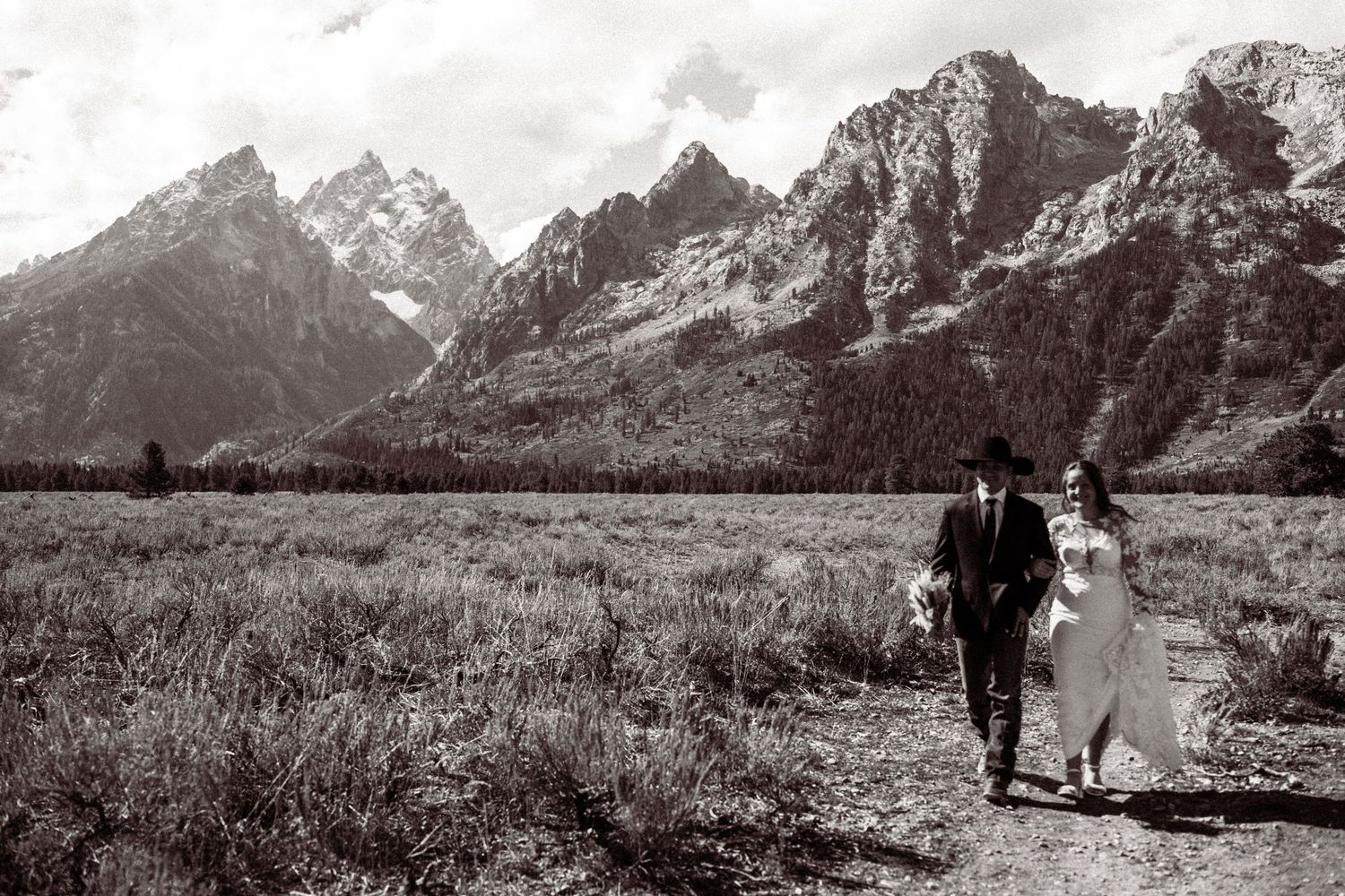 A couple walks hand in hand through a meadow with the majestic Grand Teton mountains rising dramatically in the background.