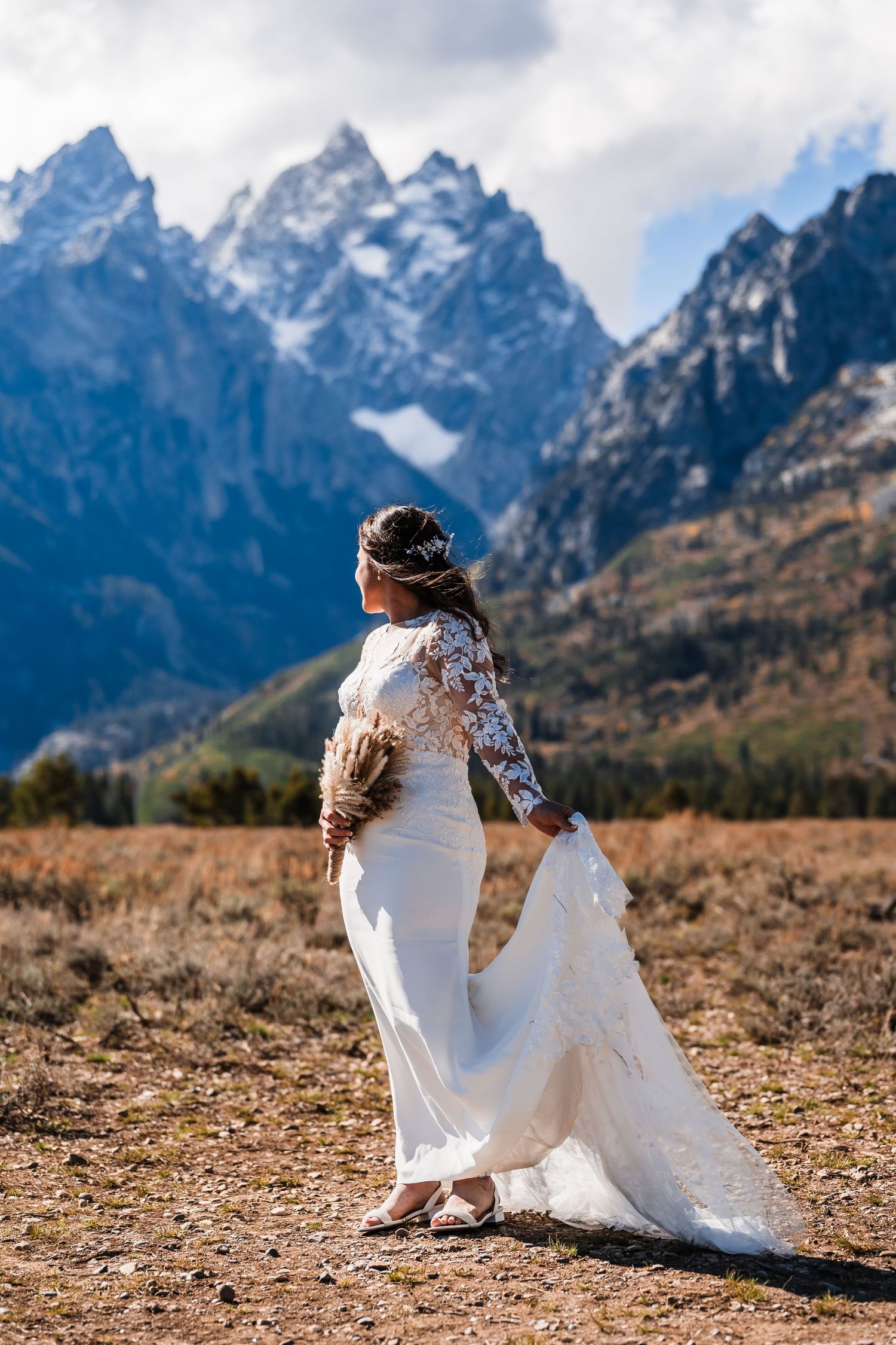 A bride in a white dress sits on the ground against a backdrop of majestic Grand Teton mountains.