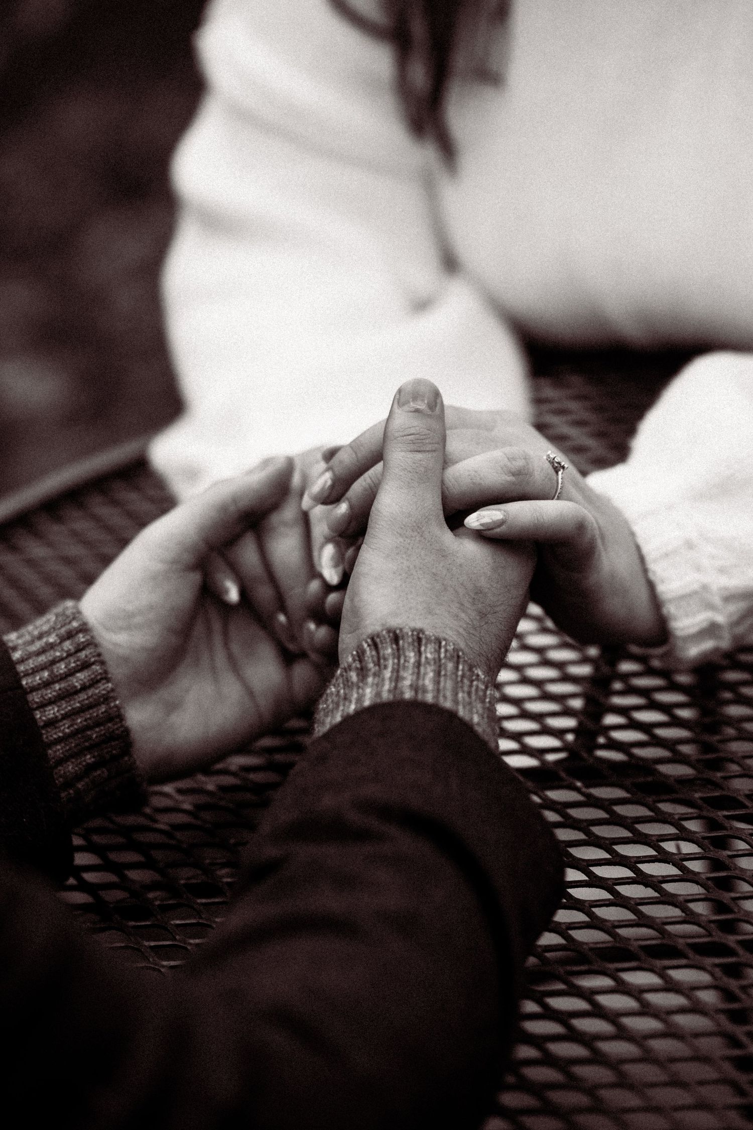 Close-up black and white photo of two people holding hands tenderly across a table with knit sleeves visible.