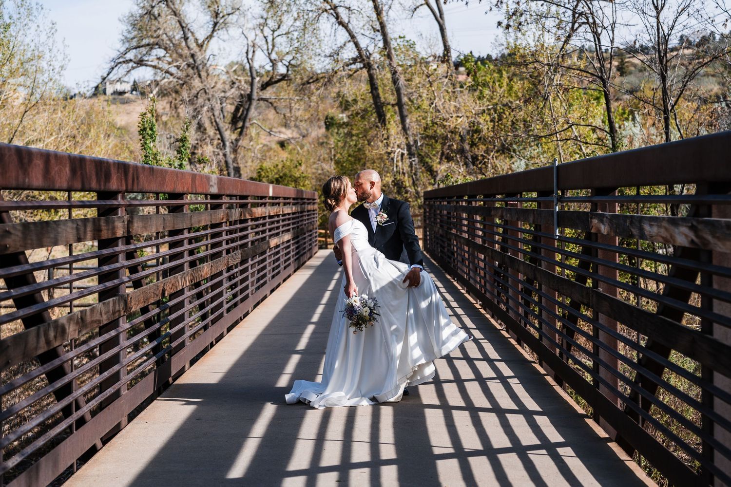 Groom lifting bride in white gown on rustic bridge surrounded by autumn trees at wedding.