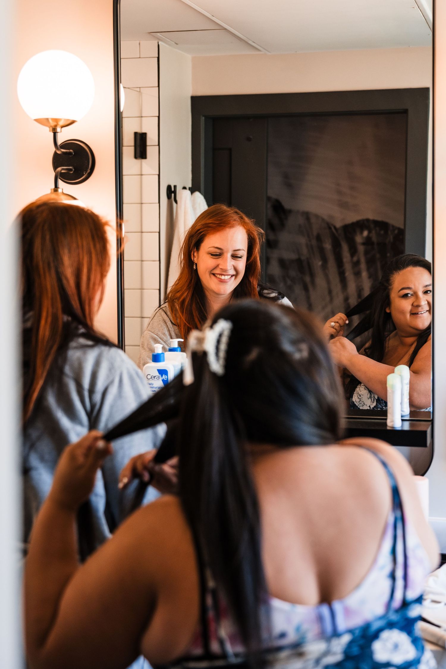 Bridesmaids laughing and styling hair together in a bathroom mirror reflection before a wedding.