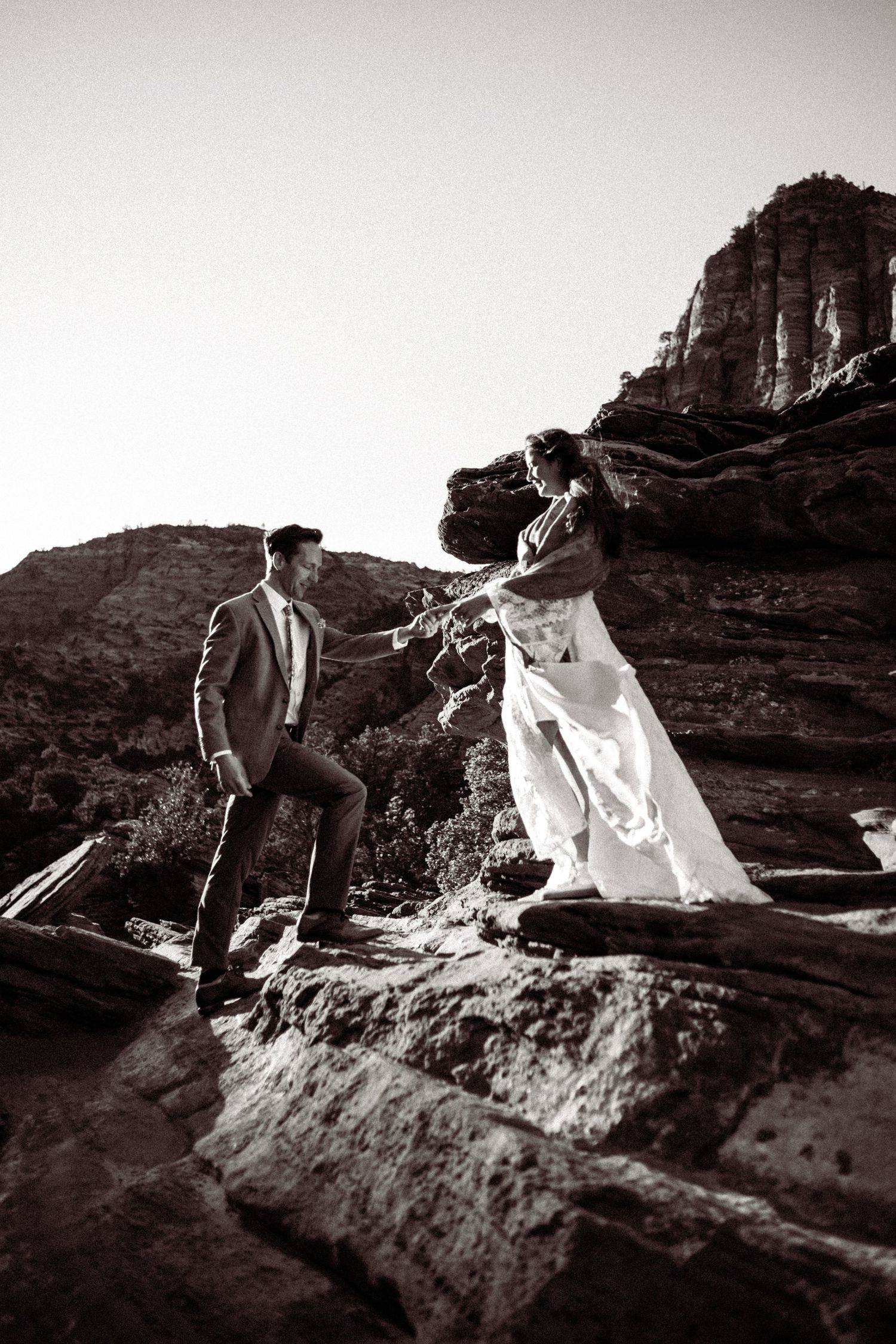 Black and white photo of bride and groom holding hands on rocky terrain with red rock formations behind them.