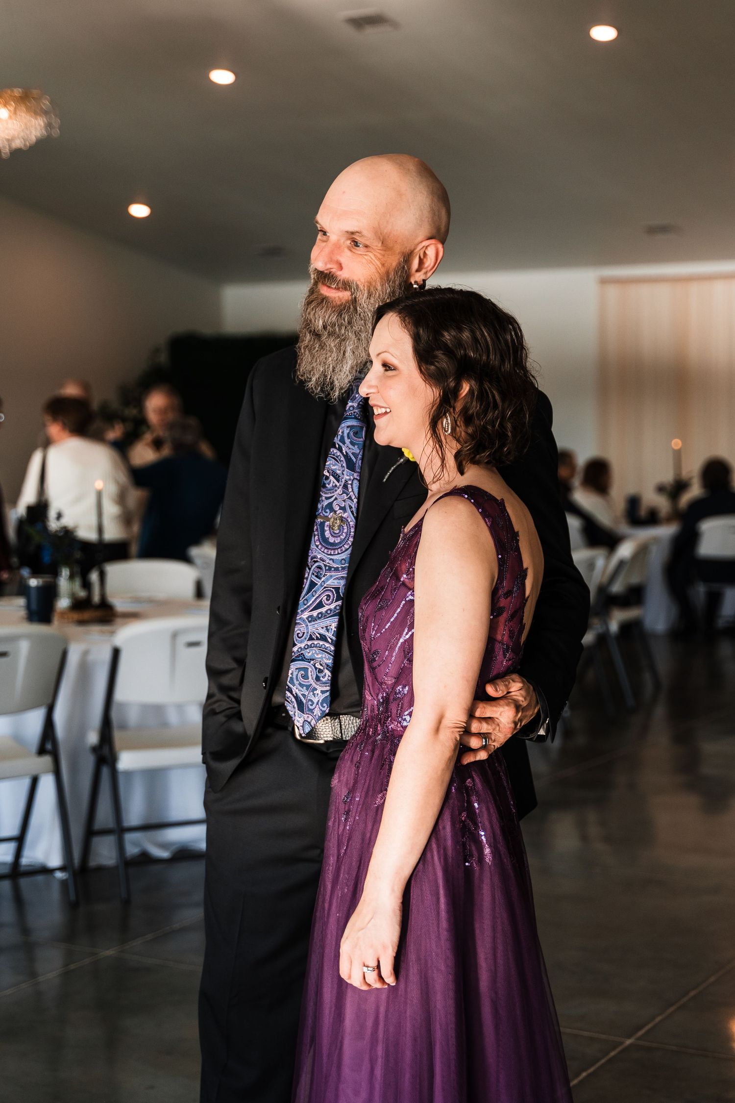 Bald groom with beard embraces bride in purple gown at wedding reception venue.