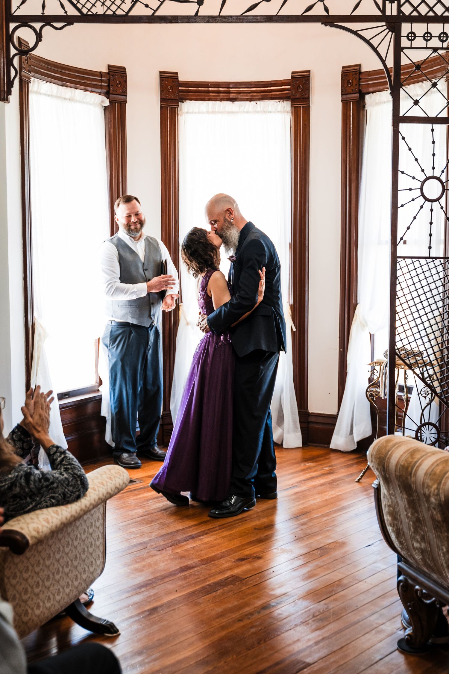Couple shares first kiss in purple wedding dress and suit in elegant Victorian home with wood floors.