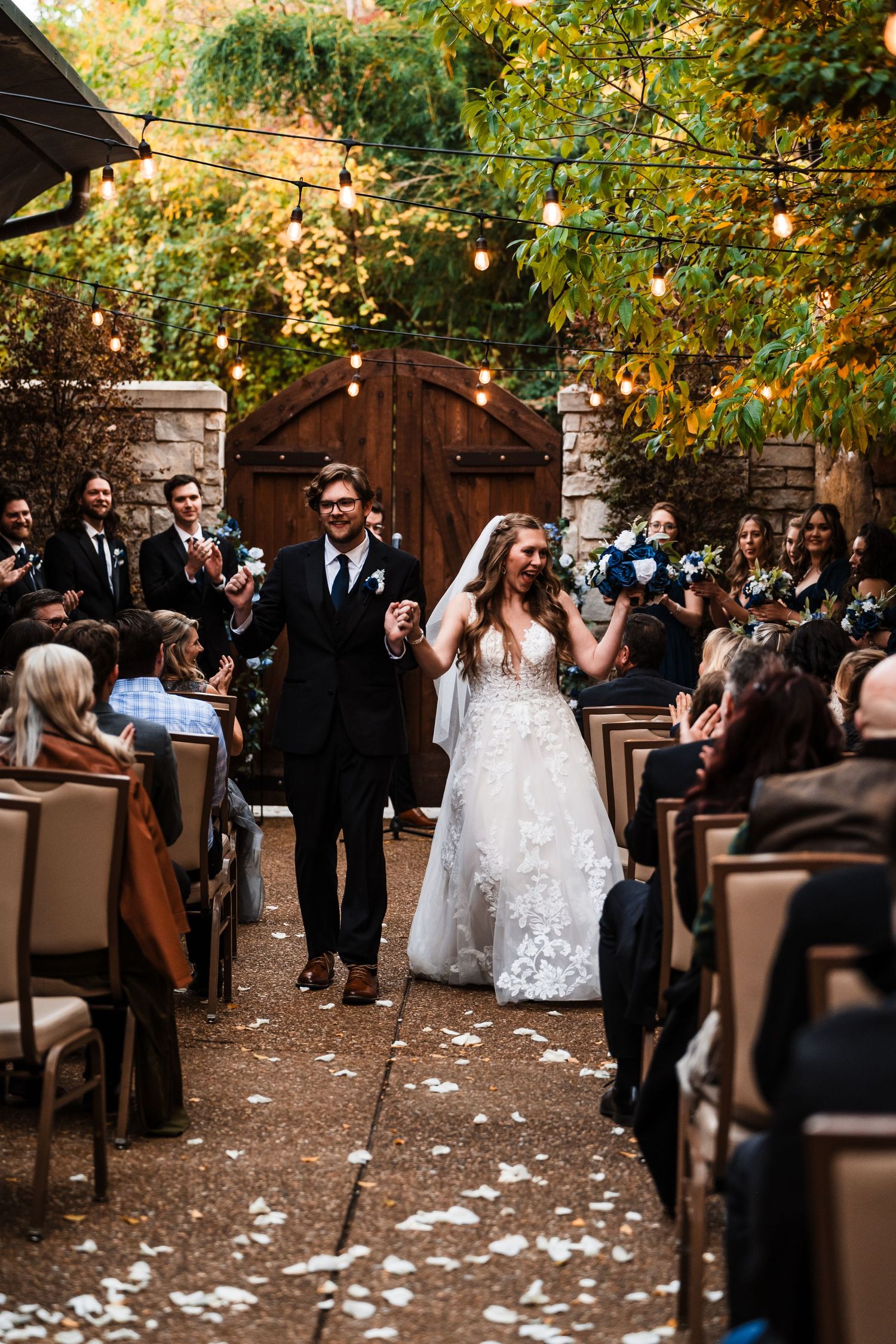 Newlyweds walk down the aisle while guests seated on brown chairs celebrate.