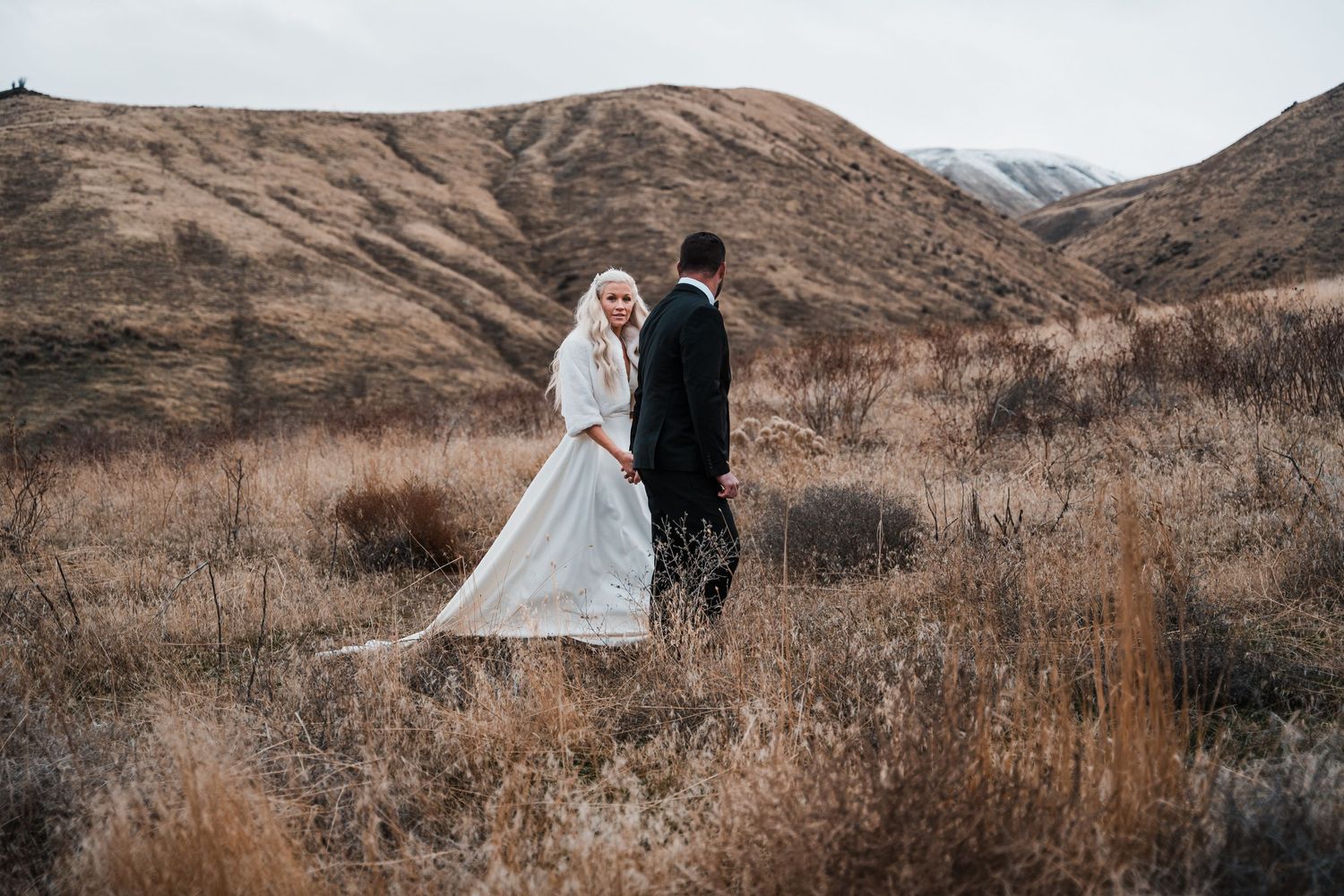 Wedding couple standing in brown grass with mountains in background.
