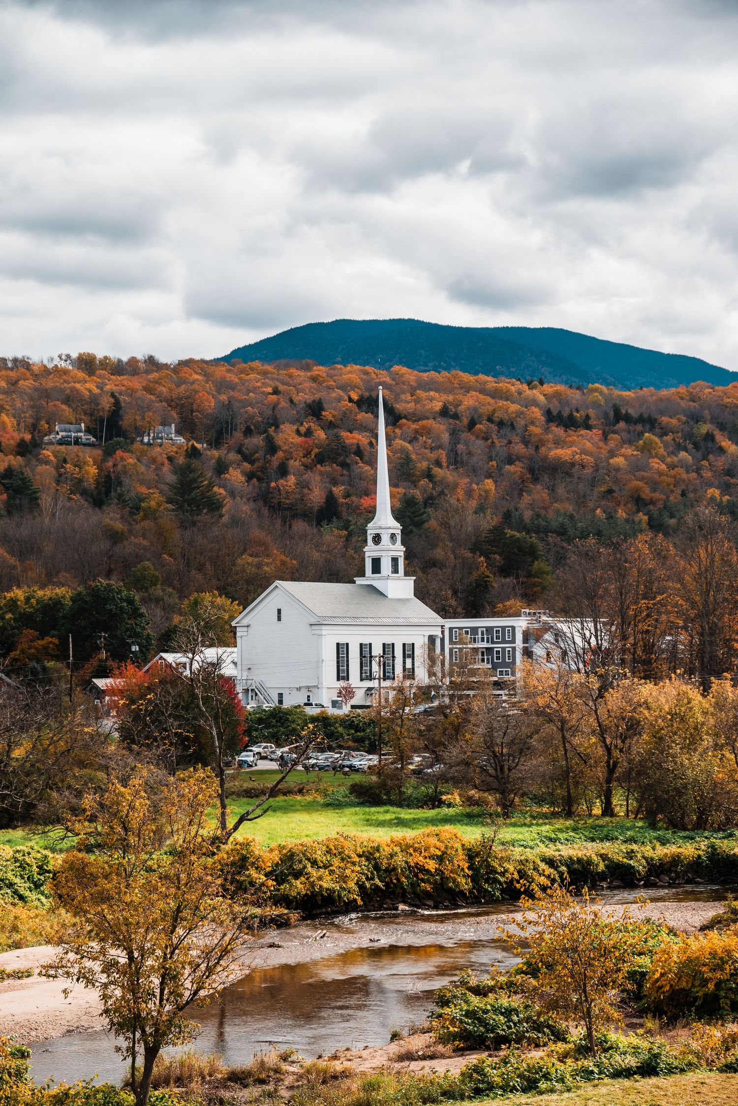 A white church with a tall steeple stands against autumn foliage and mountains in Stowe, Vermont near a winding river.