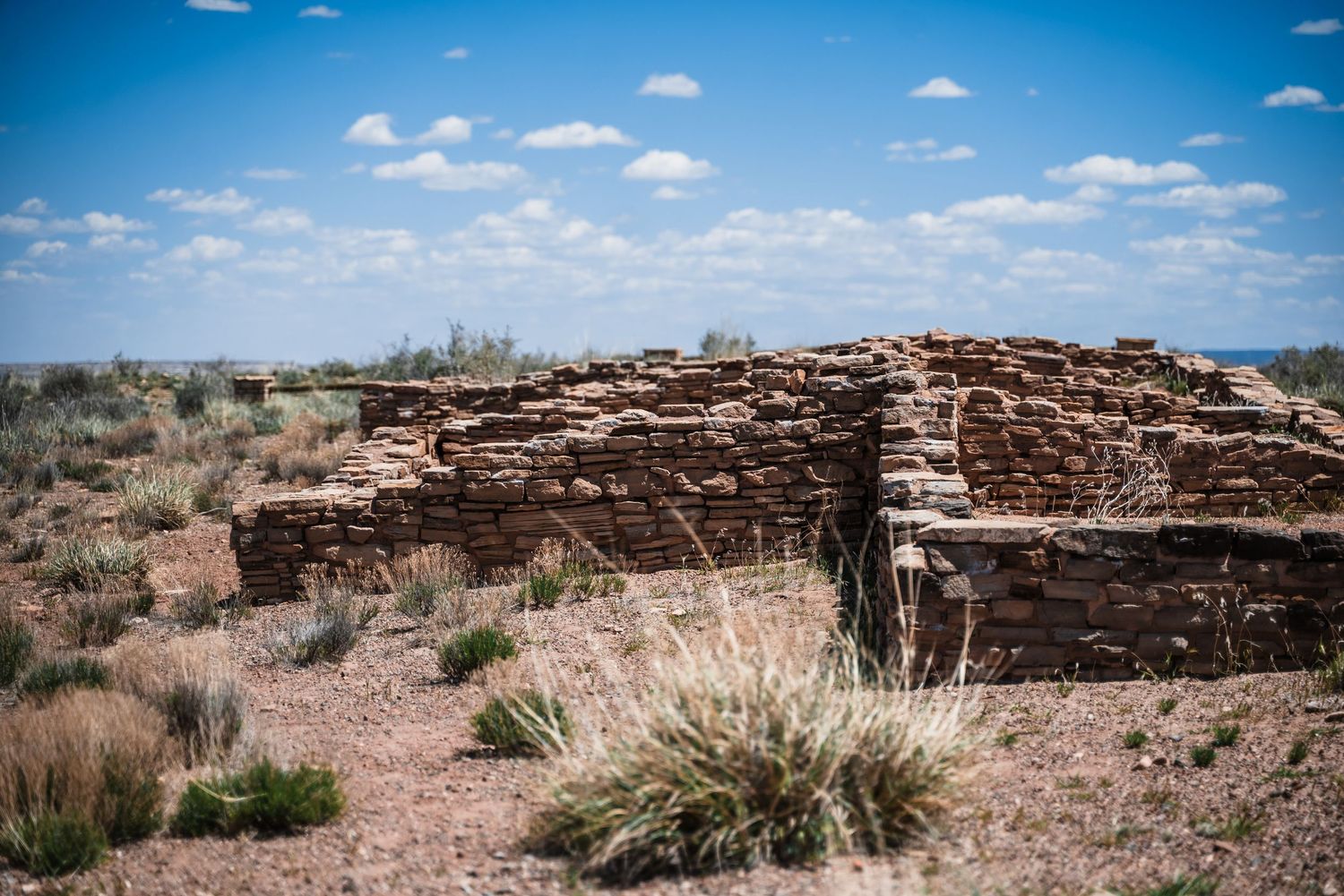 Ancient stone ruins stand in the desert landscape under bright blue skies and puffy white clouds.