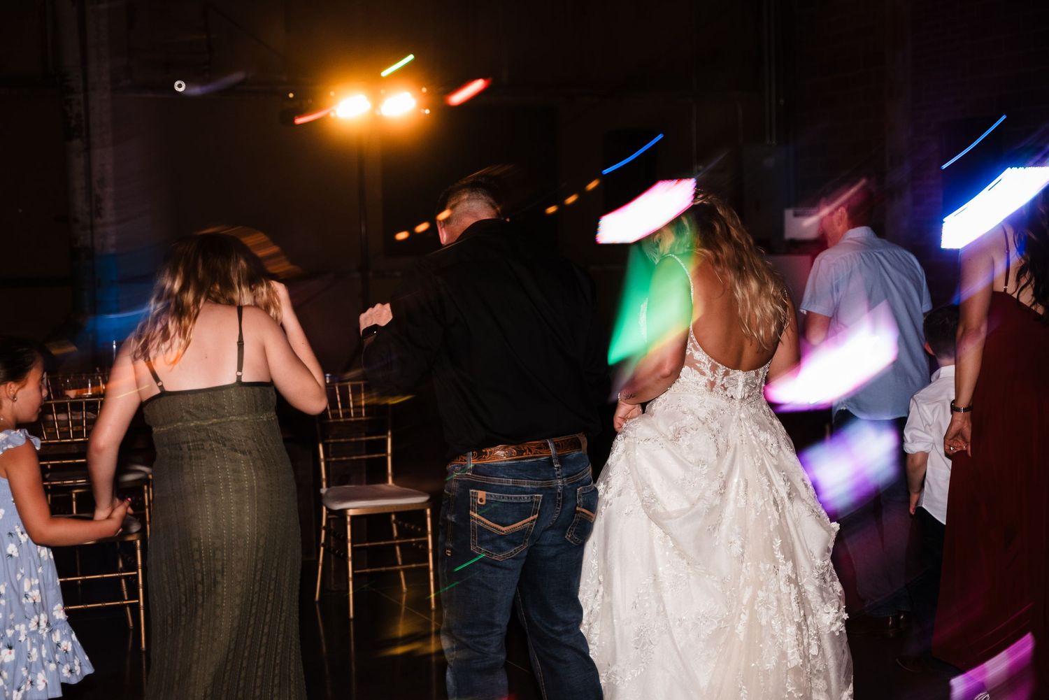 Wedding guests dance and celebrate on a dimly lit dance floor with colorful lights.
