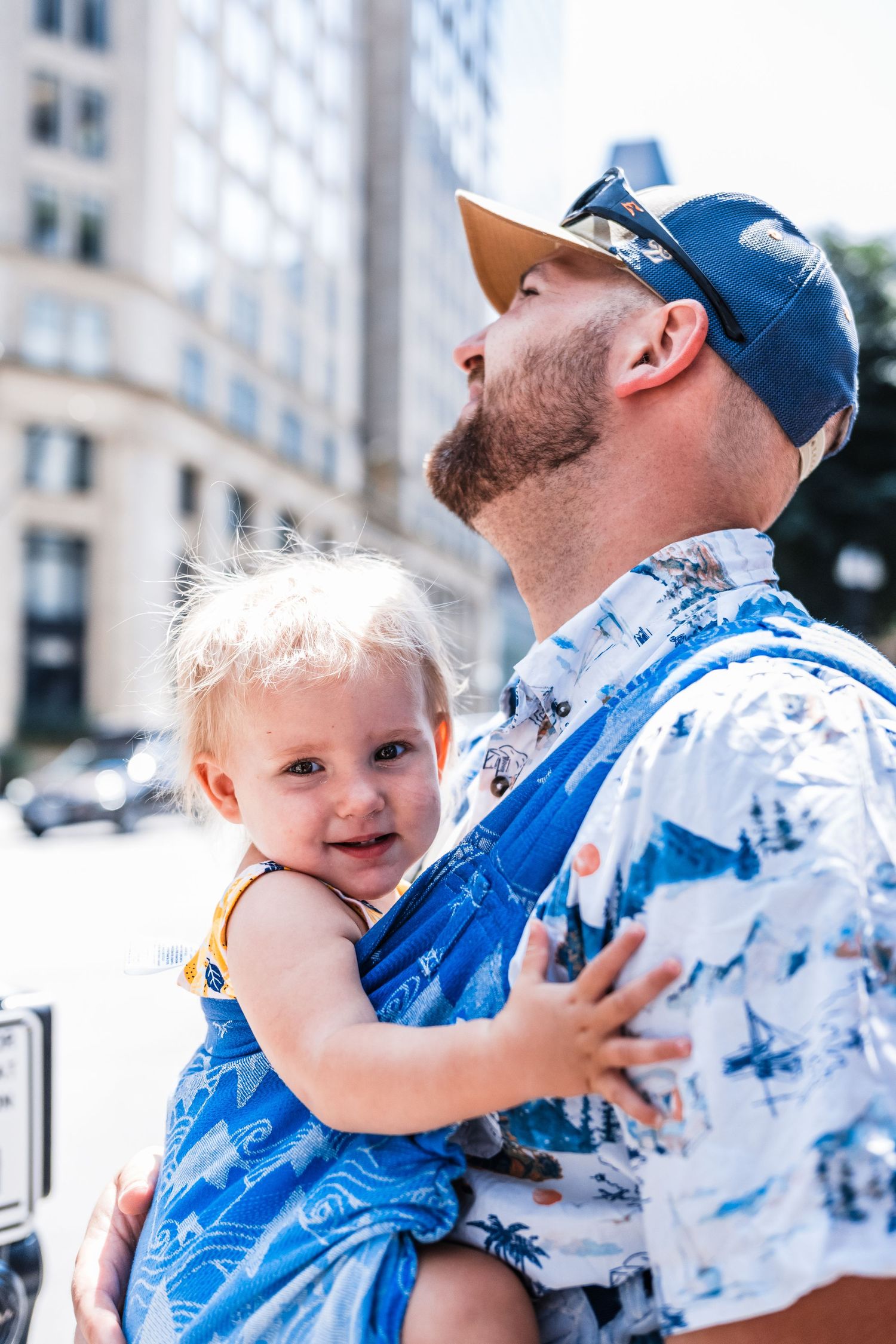 Baby wearing blue outfit smiles while being carried in city setting on sunny day.