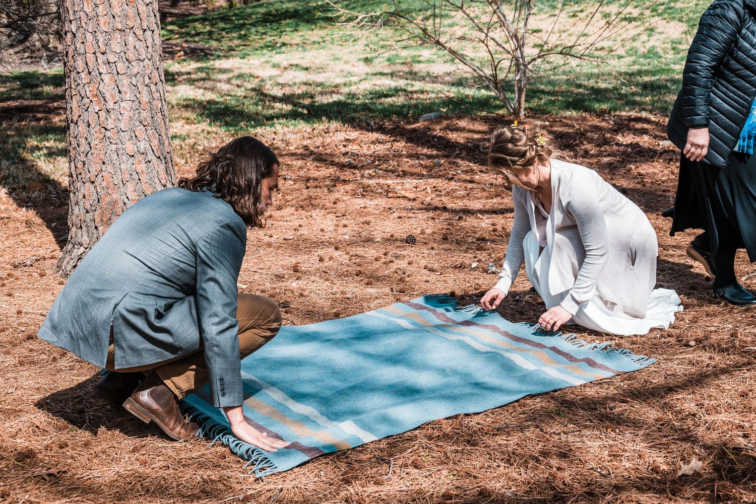 Setting up a blue blanket on the ground beneath pine trees for an outdoor ceremony.