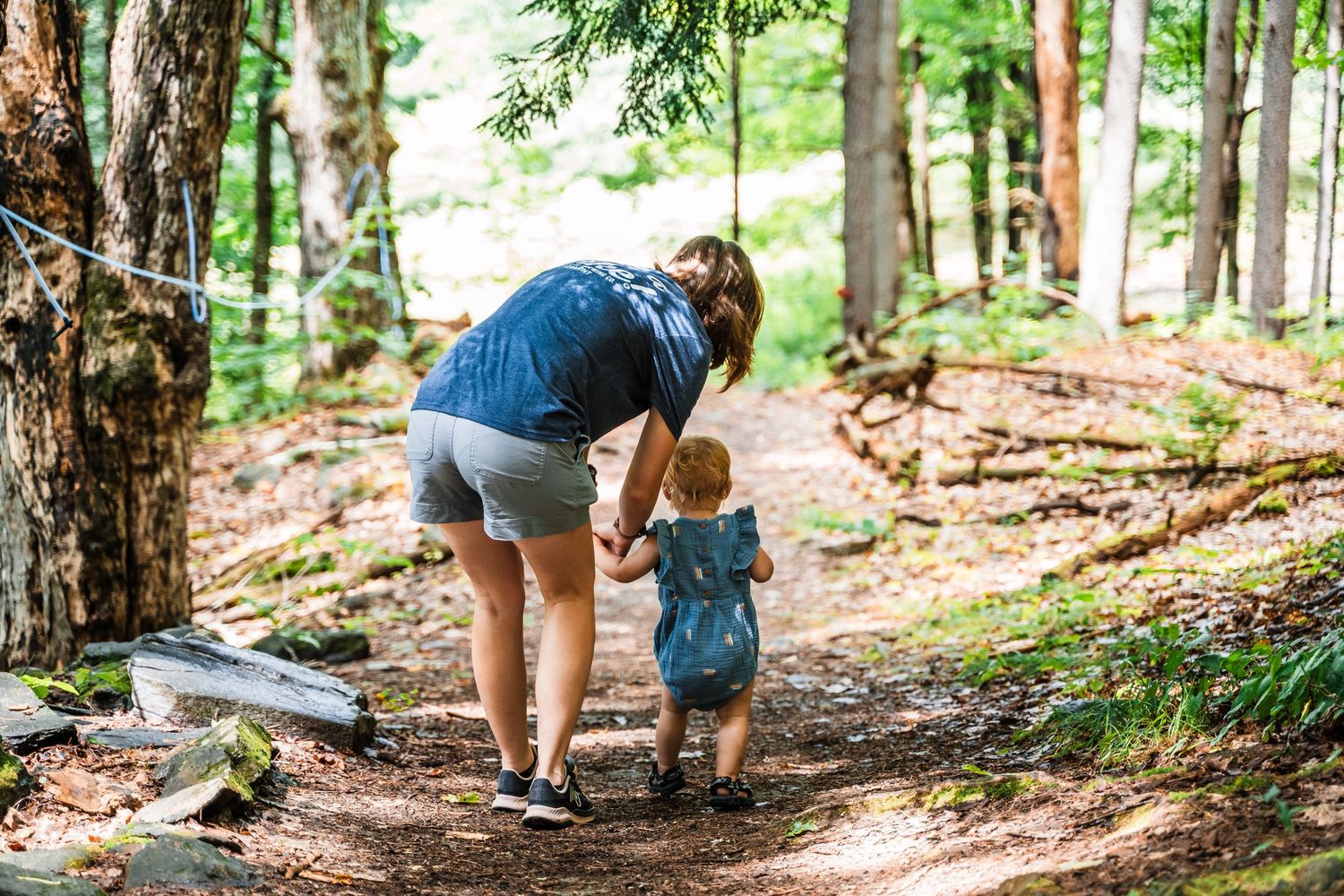 Adult and small child walking together on a woodland trail through tall trees.