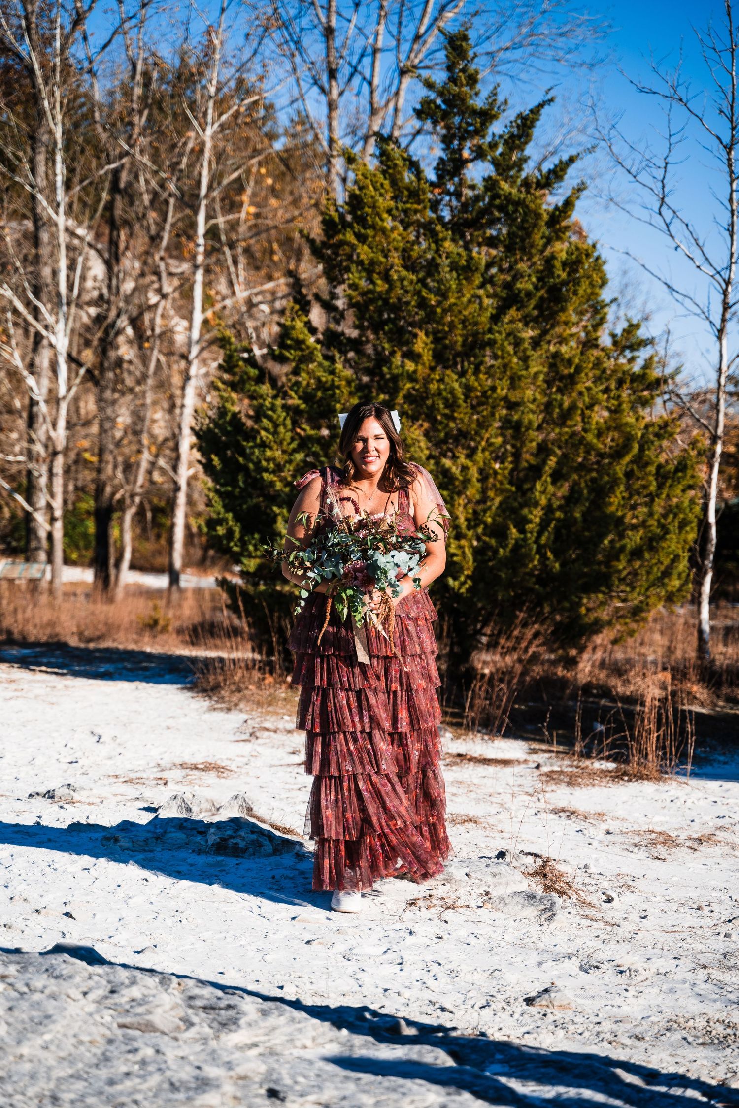 Person in a flowing burgundy dress holding a bouquet while standing in snow with evergreen trees behind.