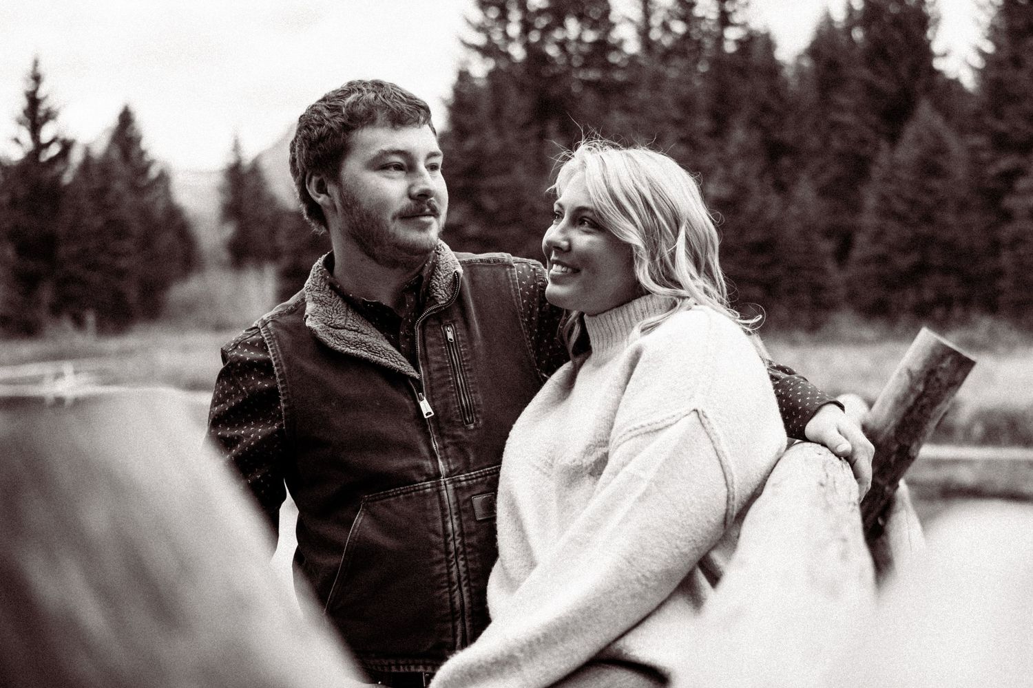 A couple shares a romantic black and white moment by a lake with evergreen trees in the background.