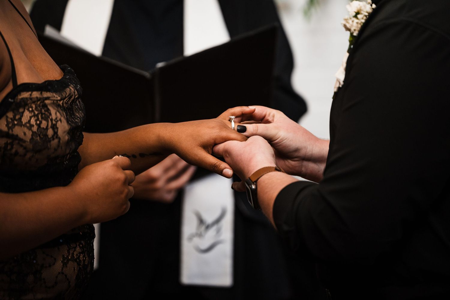 Two hands exchanging rings during an intimate wedding ceremony with an officiant in the background.