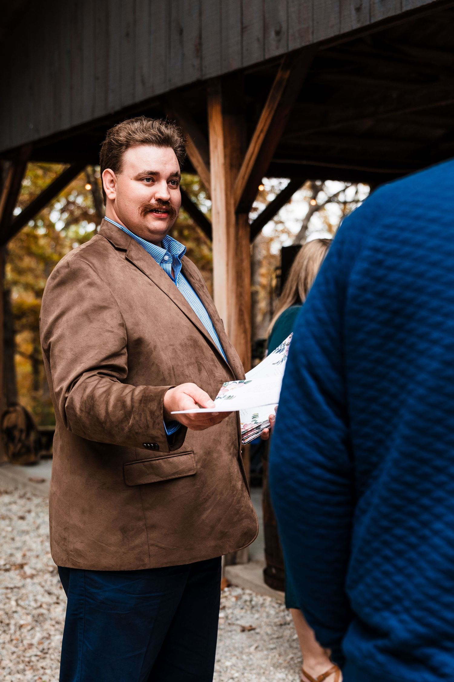 A person in a brown blazer holds papers during an outdoor gathering.