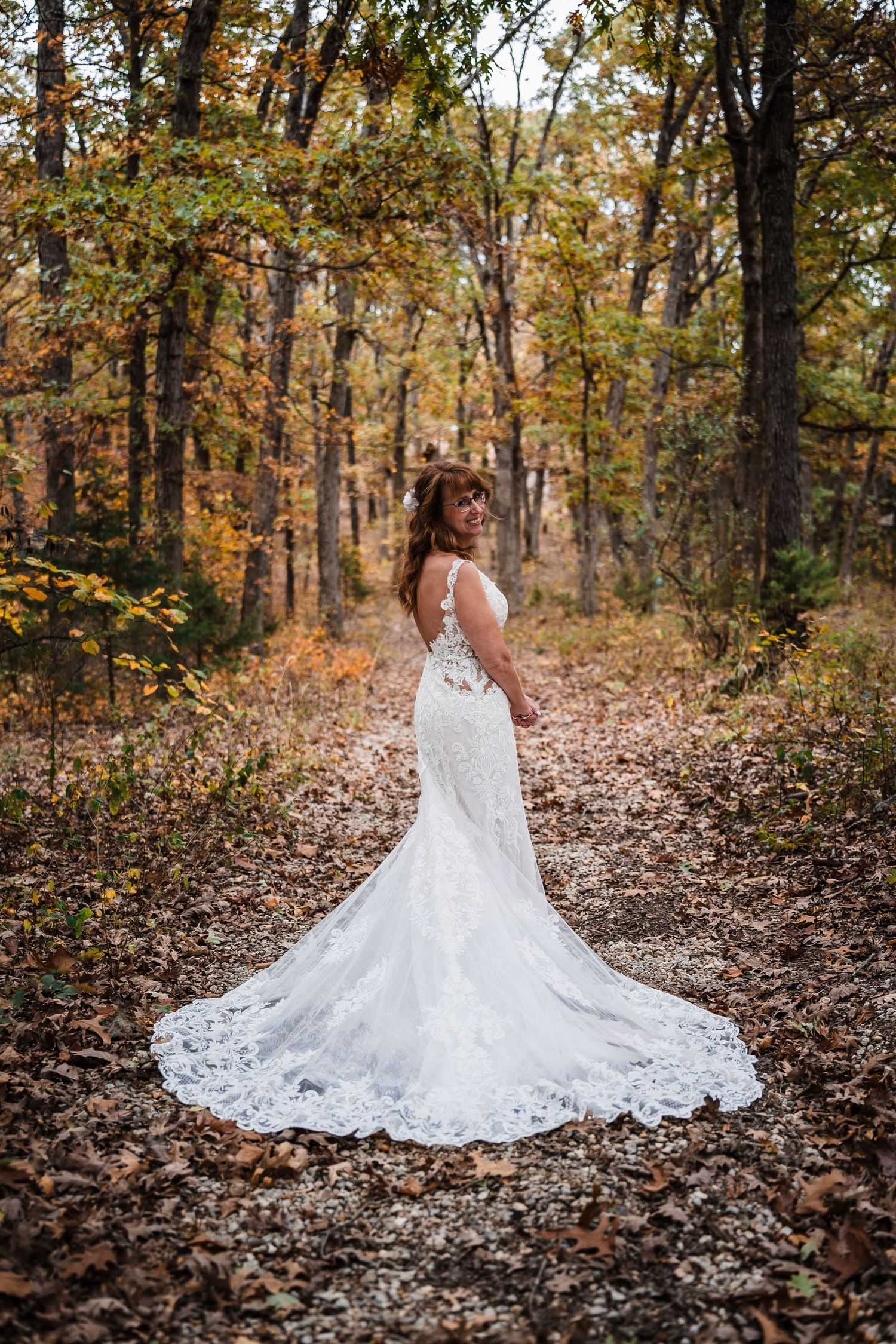 A bride in a lace wedding dress with long train poses in an autumn forest setting.