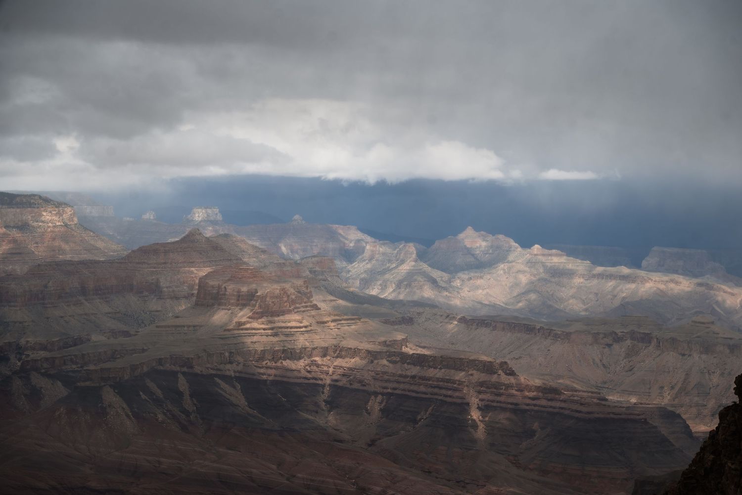 Storm clouds cast dramatic shadows over the Grand Canyon's rocky peaks and layered cliffs in golden afternoon light.
