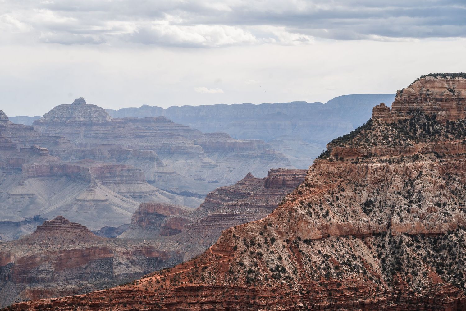 Red rock formations and distant mountain ridges stretch across the vast Grand Canyon landscape under cloudy skies.