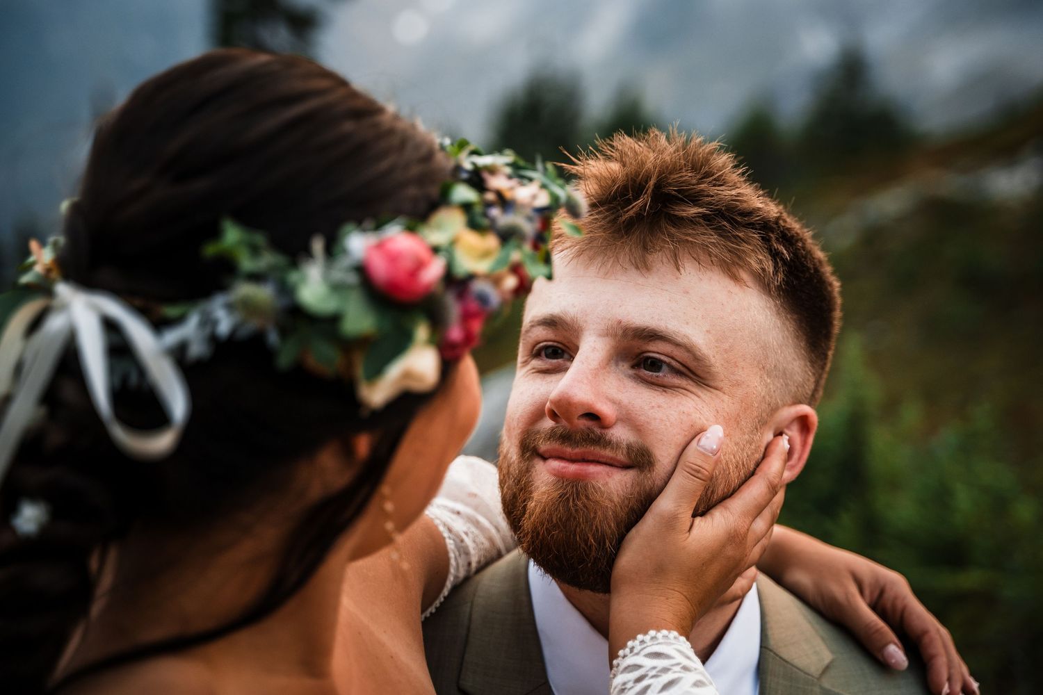 A tender moment between newlyweds outdoors, with one wearing a floral crown and the other in a suit vest.