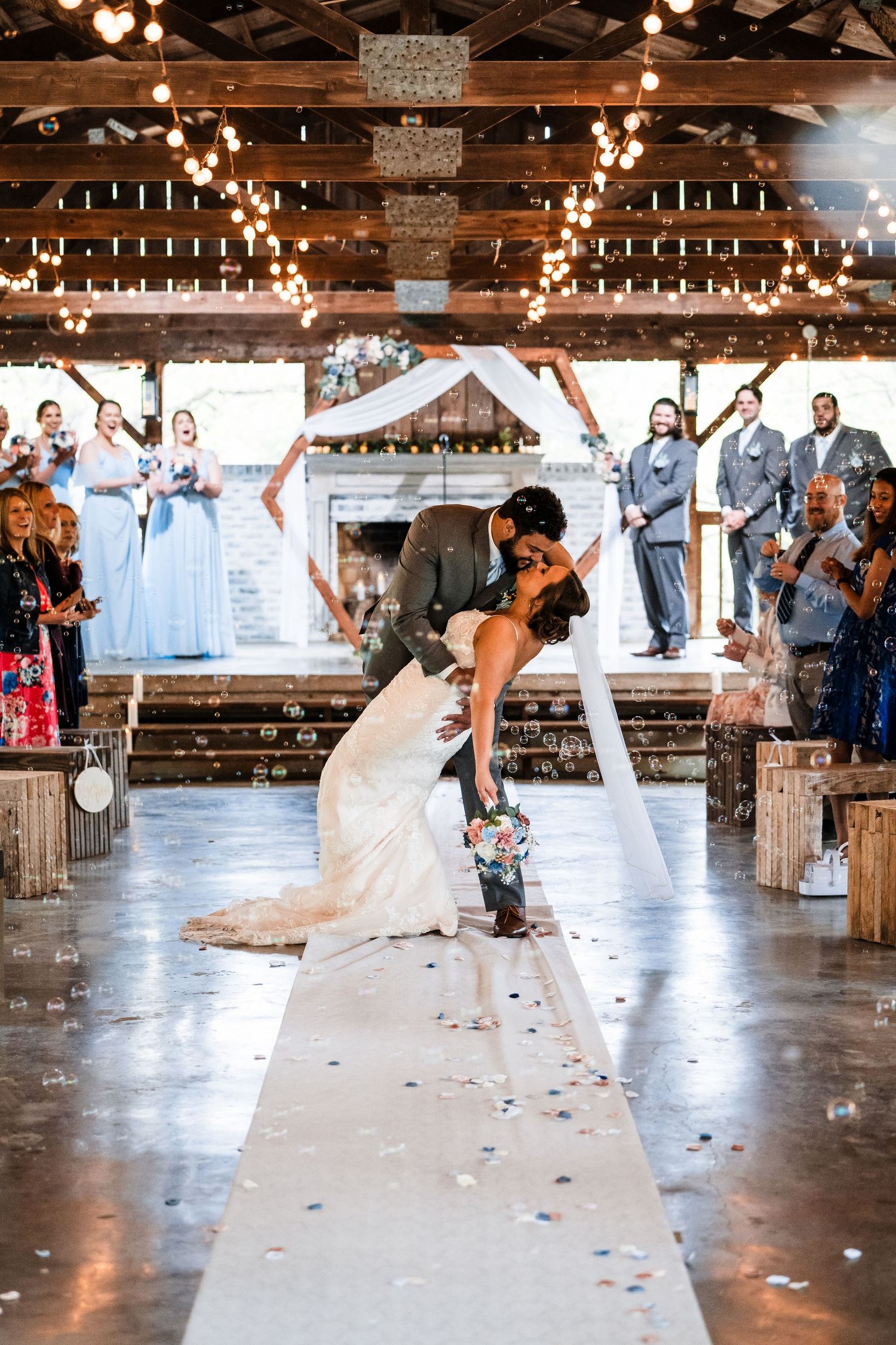 A romantic wedding ceremony in a rustic barn venue decorated with string lights and a white aisle runner.
