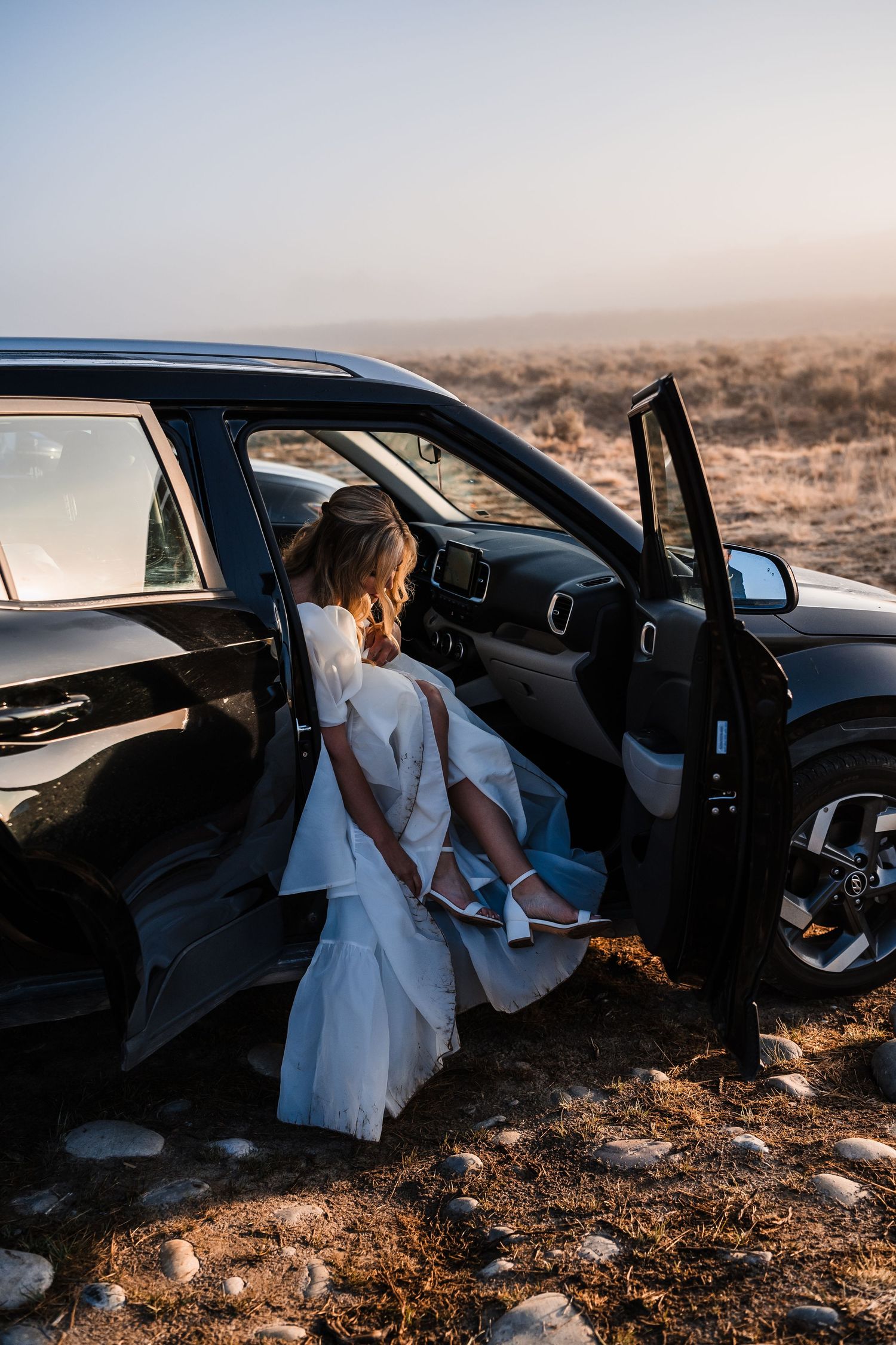 A bride in a white dress getting into a black car on a sunny day.