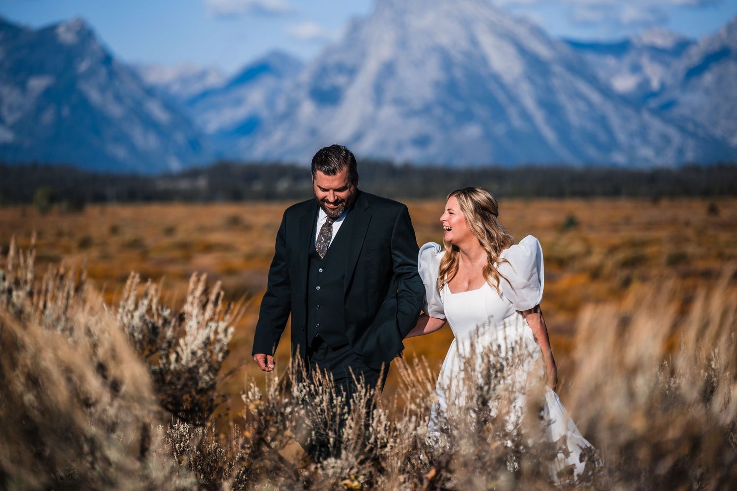 Wedding couple embraces in tall grass with dramatic mountain landscape in the background.