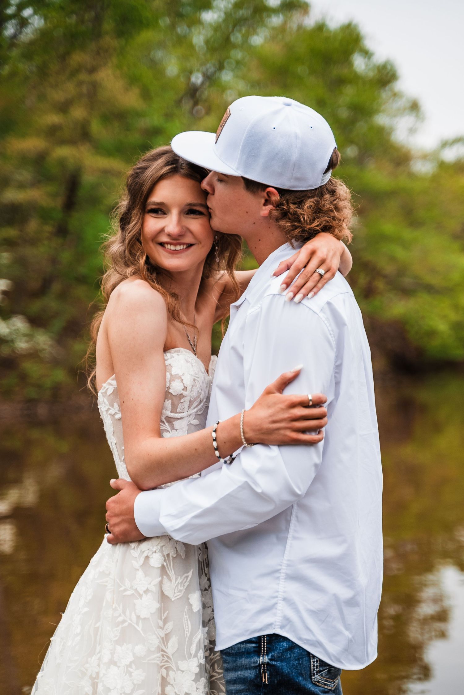 A romantic wedding photo of a couple embracing outdoors by a lake with the groom wearing a white baseball cap.
