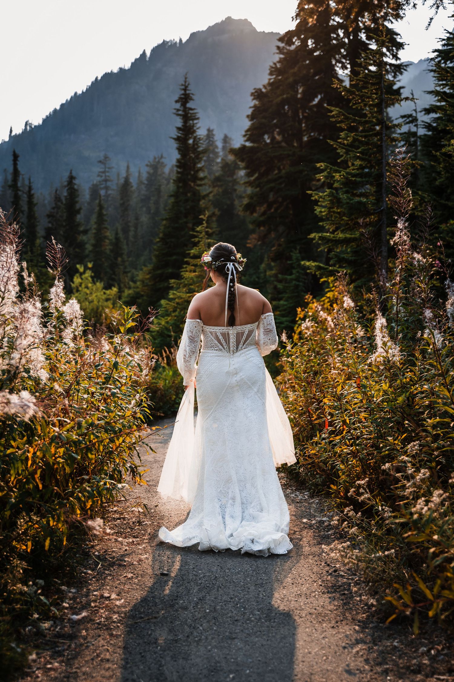 White wedding dress trails along mountain path with evergreen forest and peaks in background.