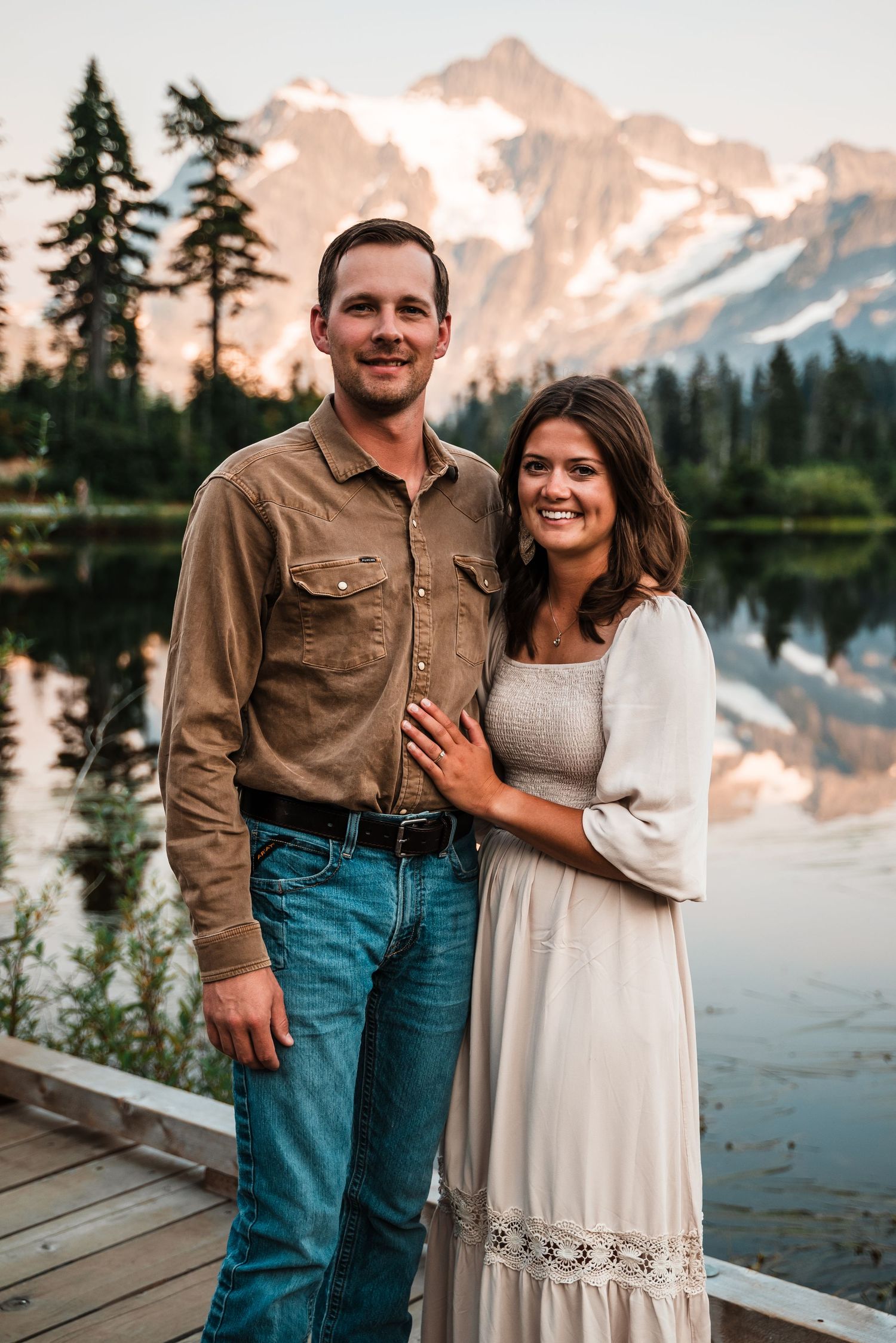 A couple in casual western attire poses by a serene mountain lake at sunset, with evergreen trees in the background.