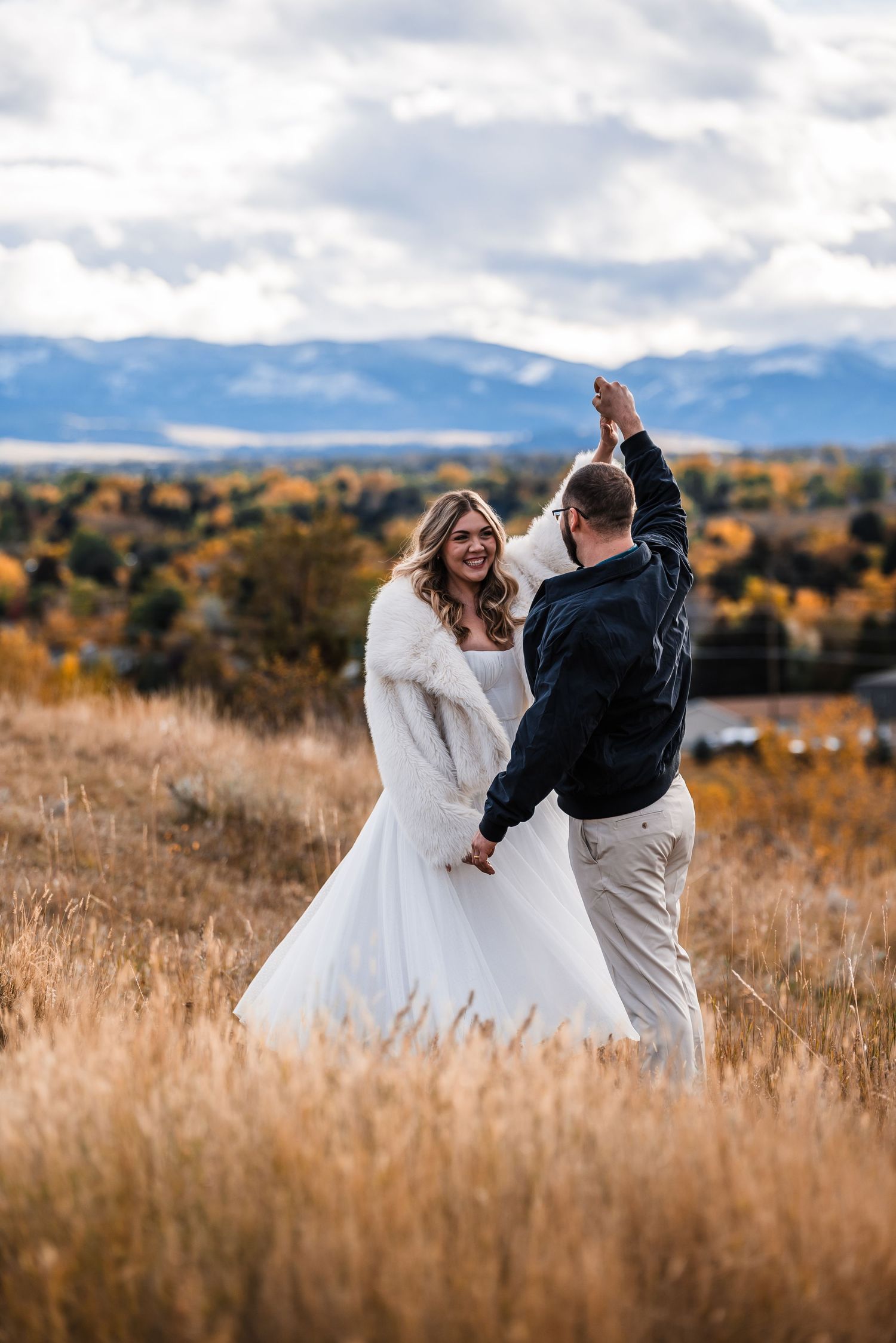 A couple in wedding attire dances together in a golden field with mountains and autumn foliage in the background.