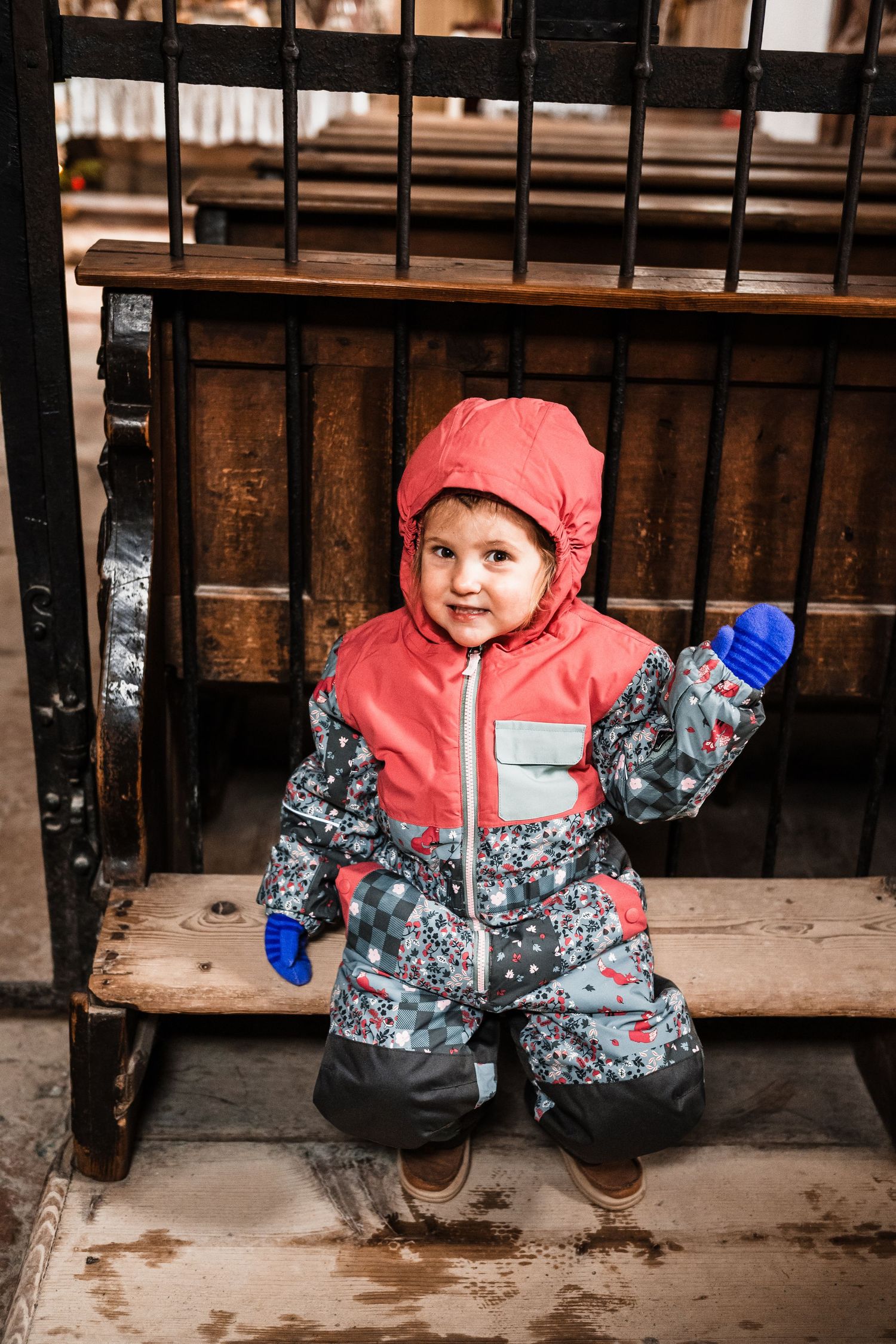 Child in pink hooded winter coat and patterned snowsuit sits on a wooden bench in snowy weather.