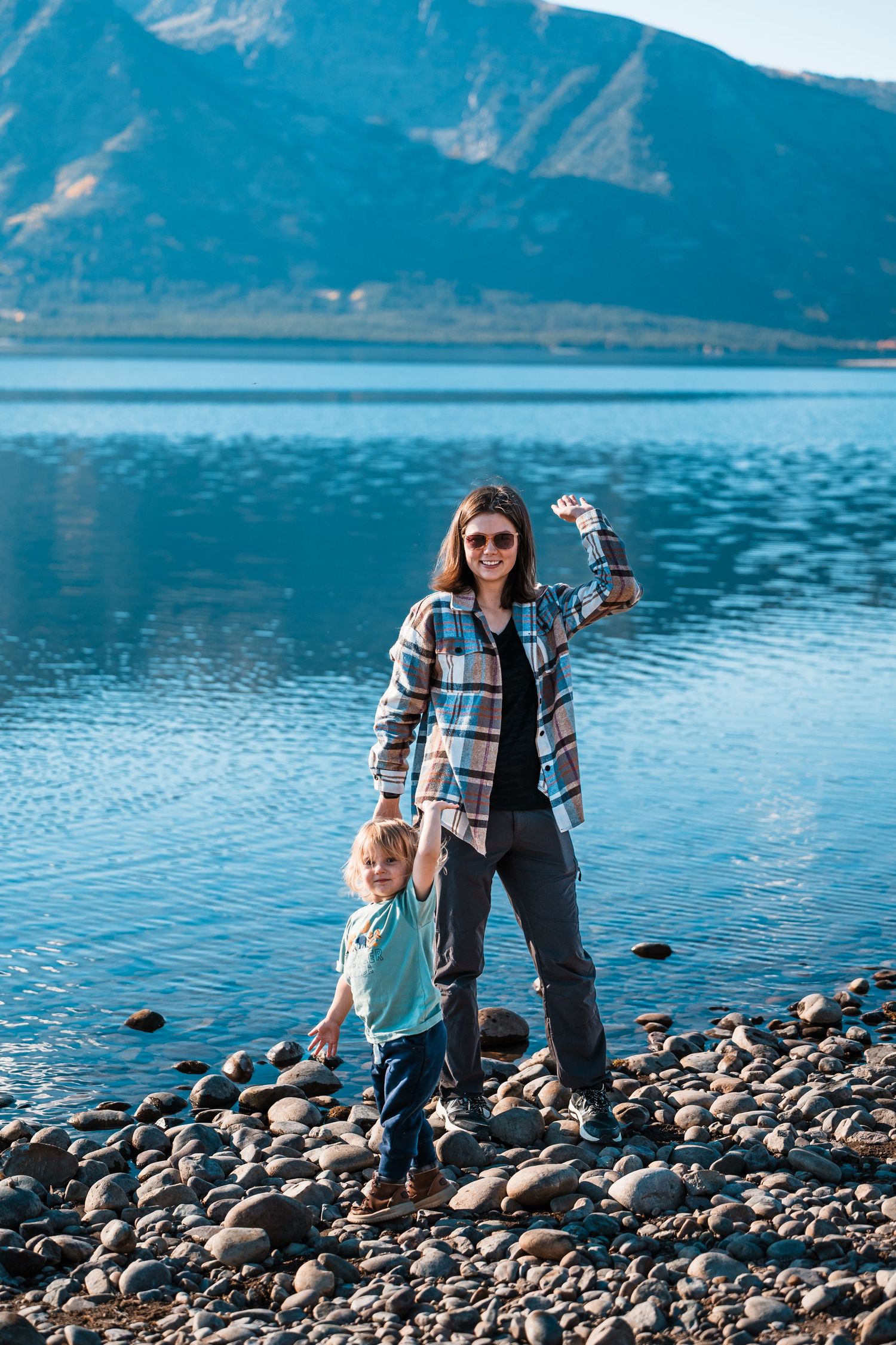 Family enjoying a peaceful moment on a rocky beach shore with mountains and blue lake in the background.