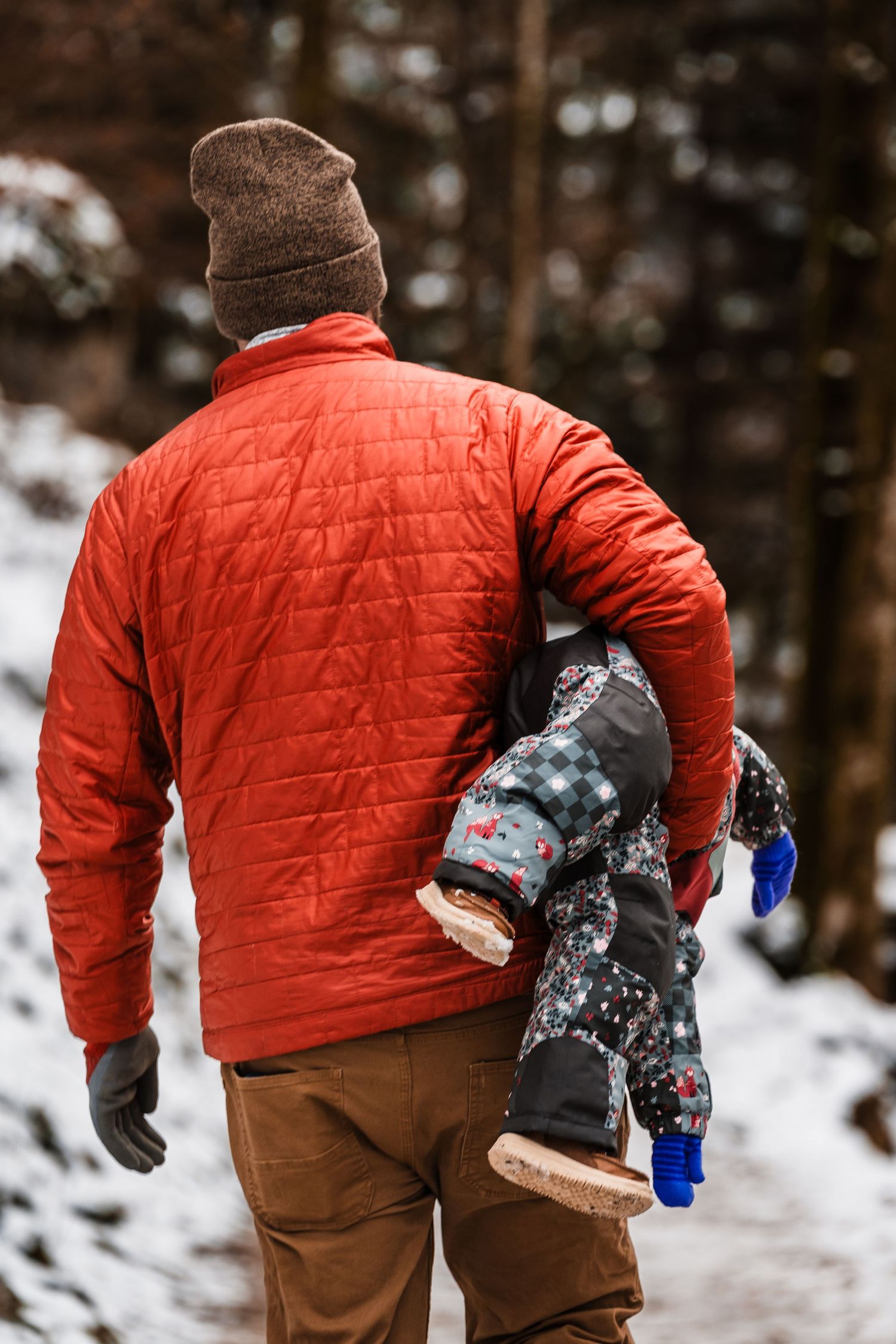 Person in bright orange quilted jacket and brown pants walking through snowy winter forest path while carrying supplies.