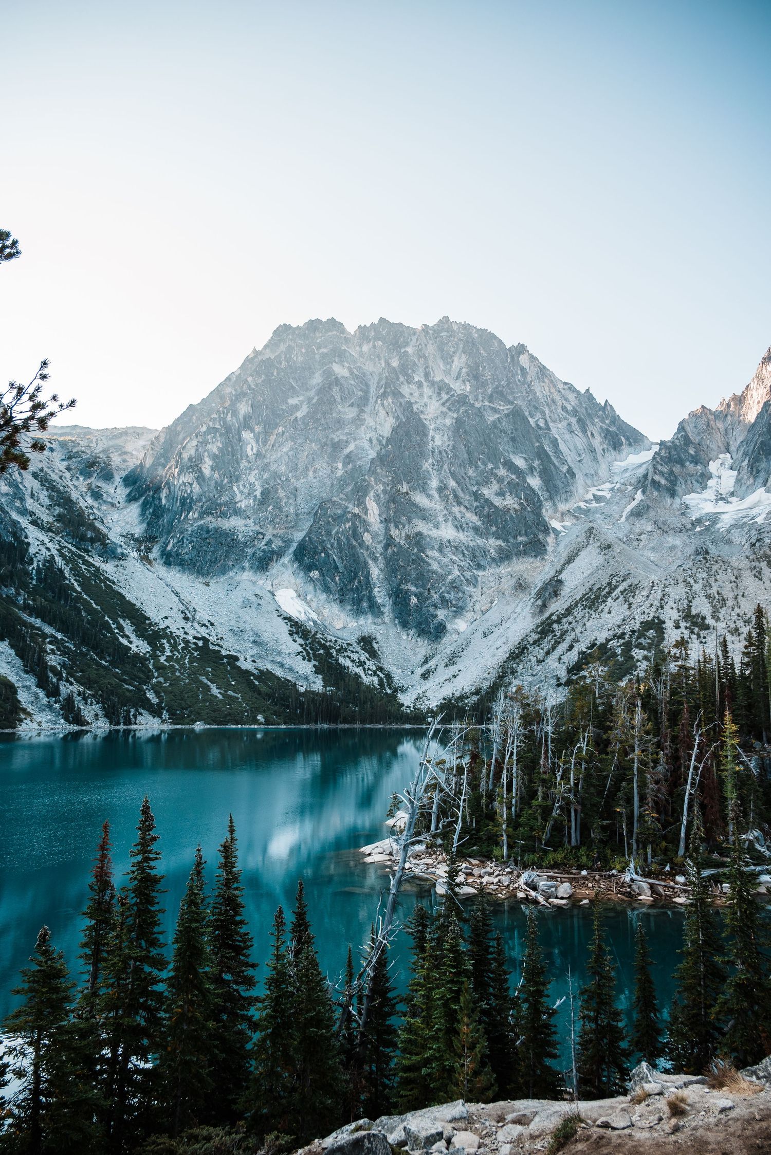 Snow-capped mountains tower over a pristine alpine lake surrounded by evergreen trees in a serene winter landscape.