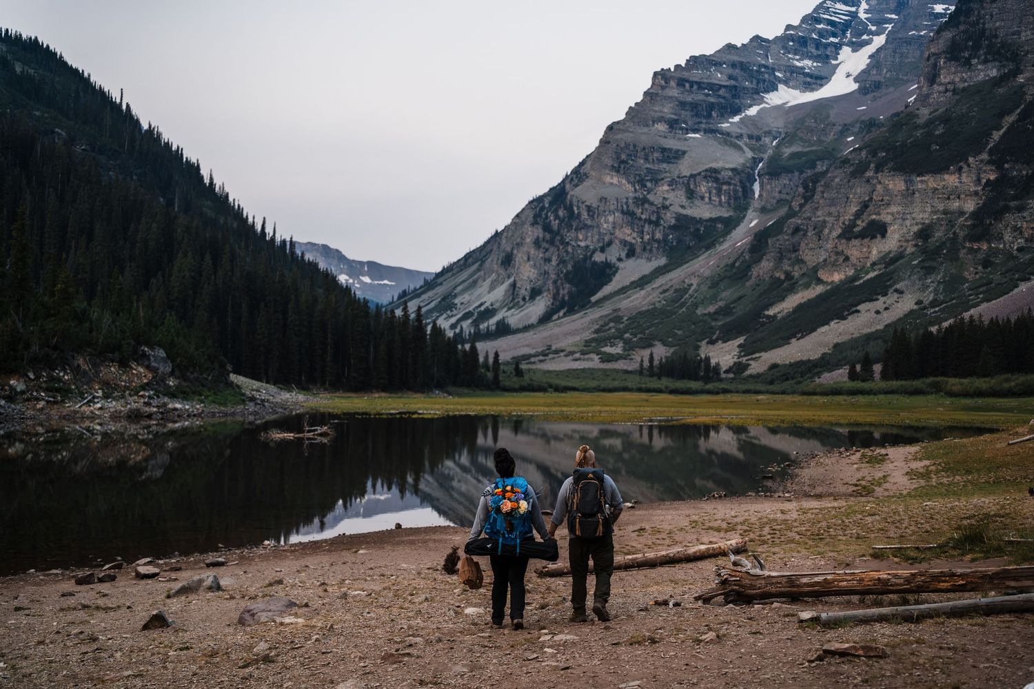 Couple holding hands by a serene mountain lake surrounded by pine trees and snow-capped peaks in the wilderness.