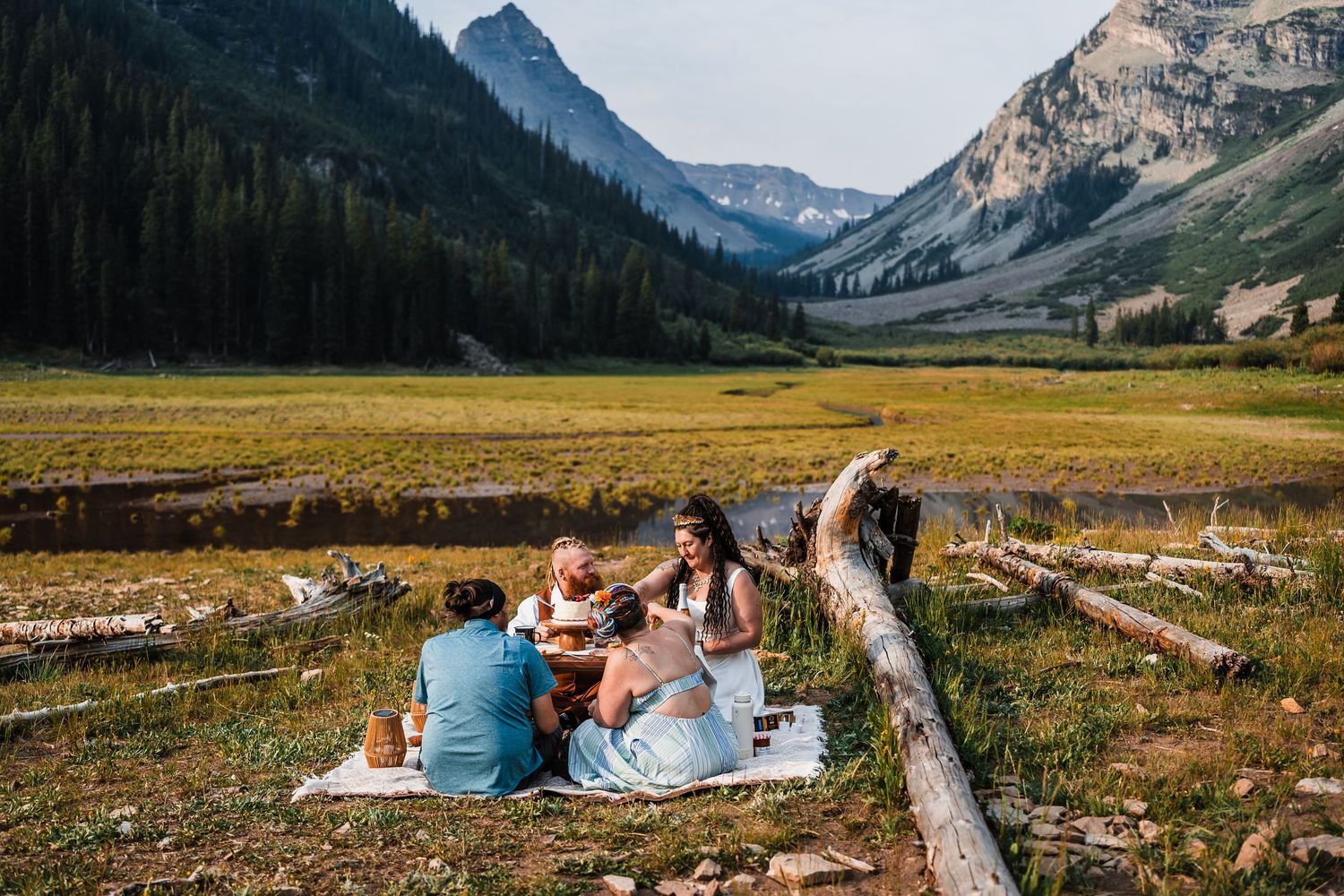 Wedding party celebrating with drinks on a picnic blanket in a scenic mountain valley meadow.
