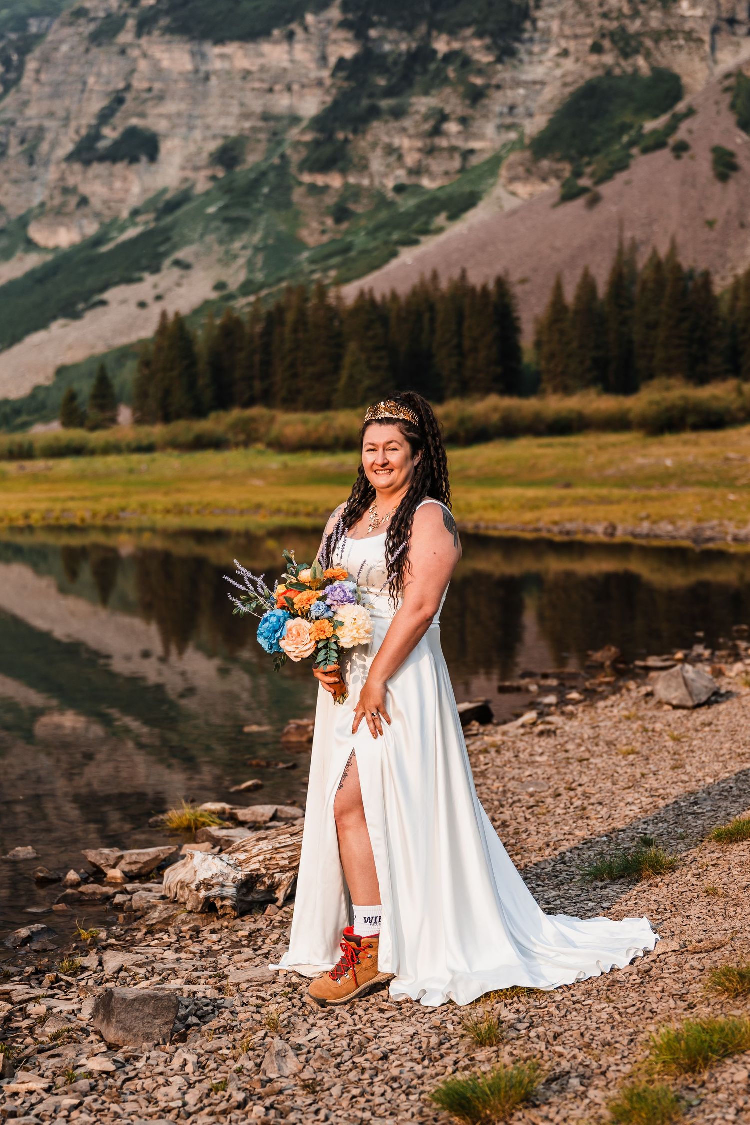 Bride in white gown with colorful bouquet stands by mountain lake at outdoor elopement ceremony.