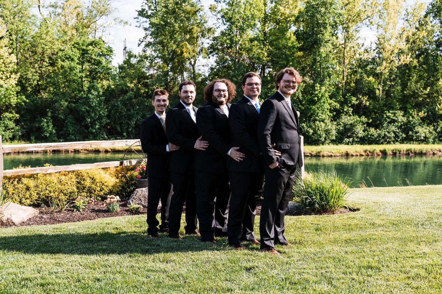 Groom and four groomsmen posing in black suits by a pond at an outdoor wedding venue.