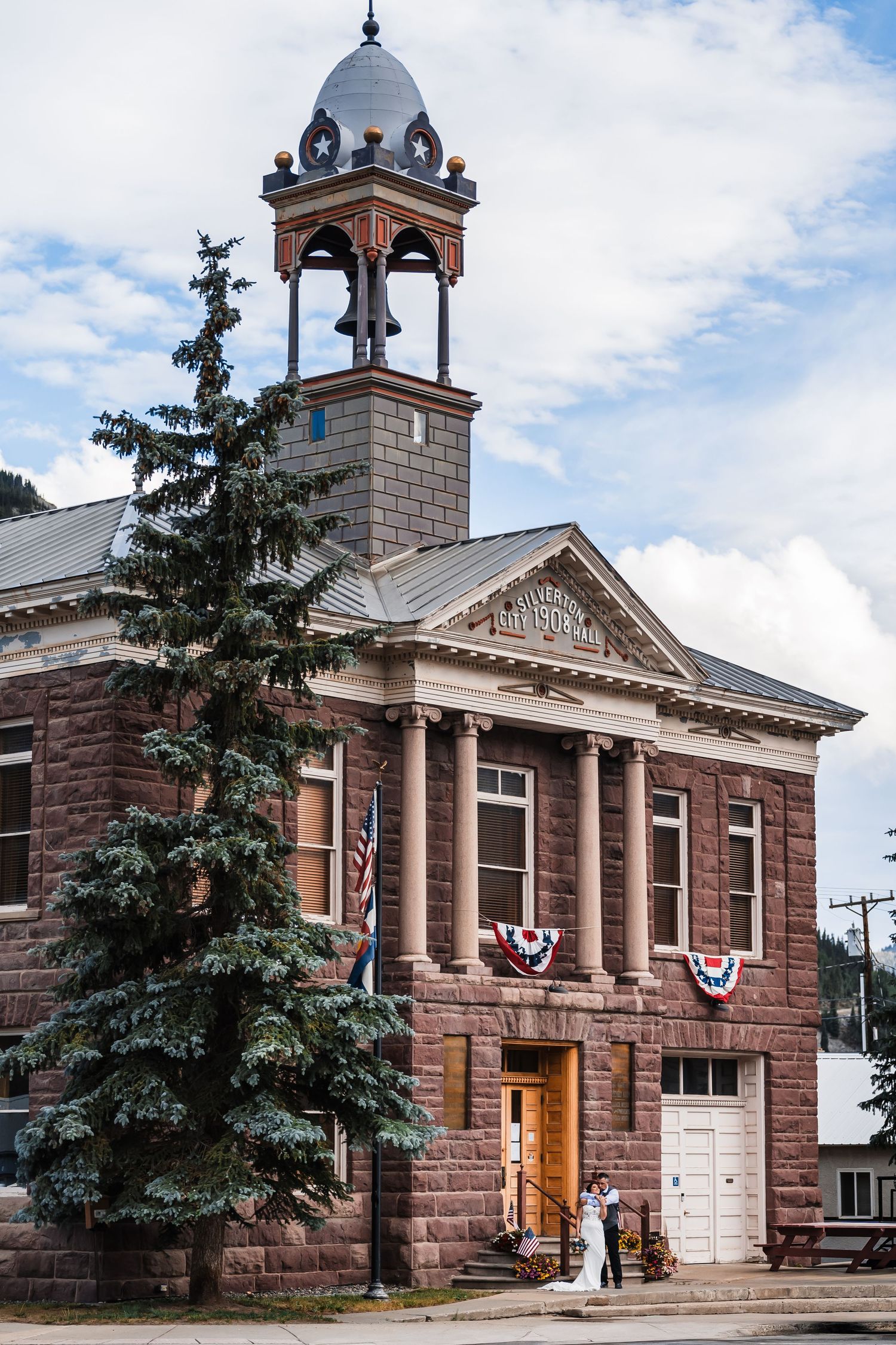 Bride and groom pose before a historic red stone courthouse decorated with American flag bunting.