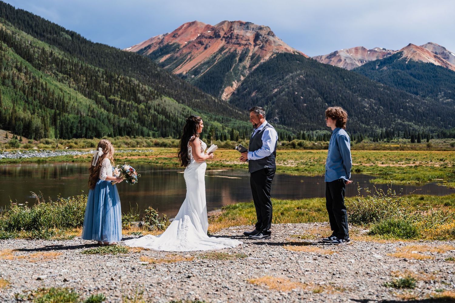 Outdoor wedding ceremony with couple exchanging vows near a mountain lake, surrounded by pine trees and red rocky peaks.