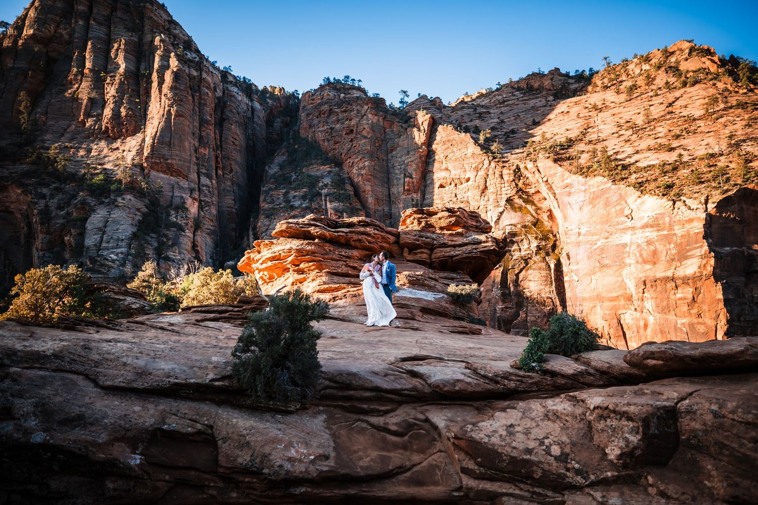 Couple in wedding attire embracing on red rock formations at Zion National Park with canyon walls behind.