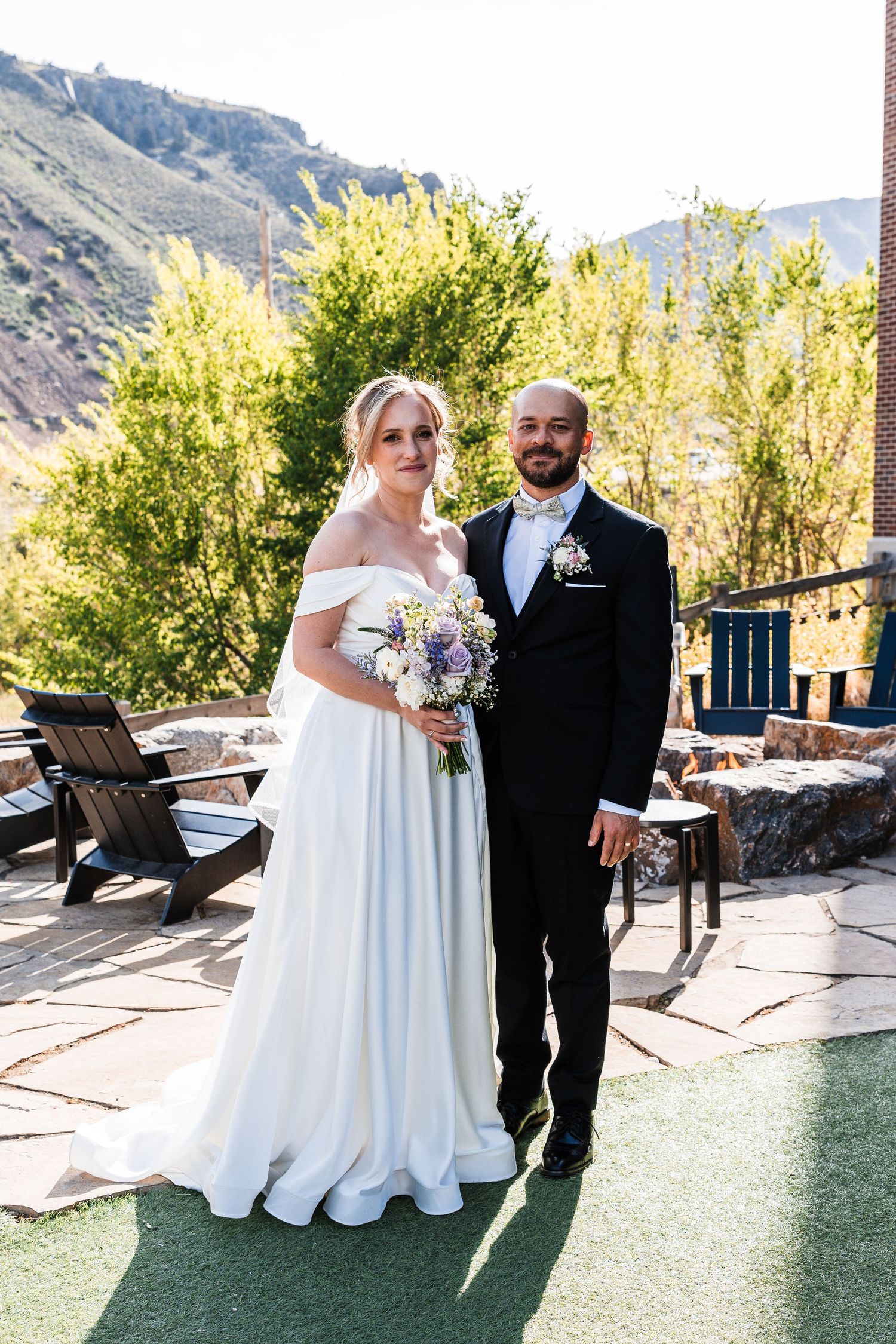 Bride in off-shoulder gown and groom in black tuxedo pose together at outdoor mountain wedding.