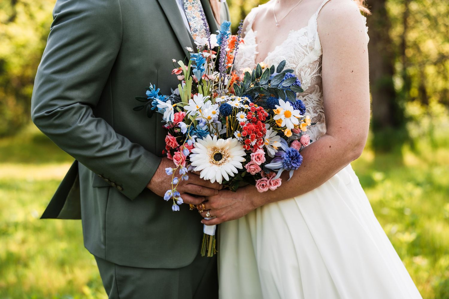 Bride and groom holding a vibrant wildflower bouquet with daisies, roses, and blue flowers outdoors.
