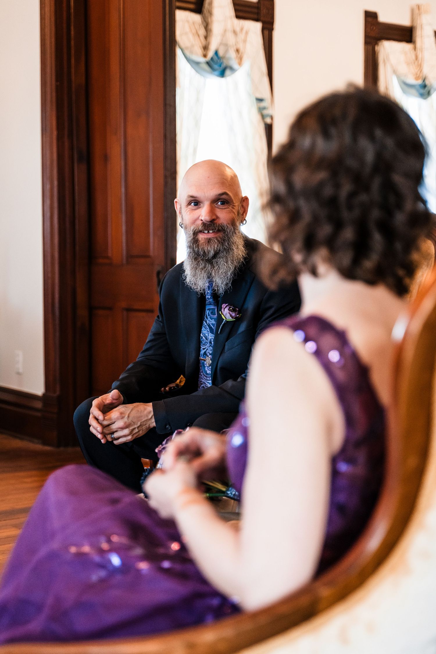 Bald bearded groom smiles at bride in purple sequin dress during intimate seated wedding ceremony indoors.