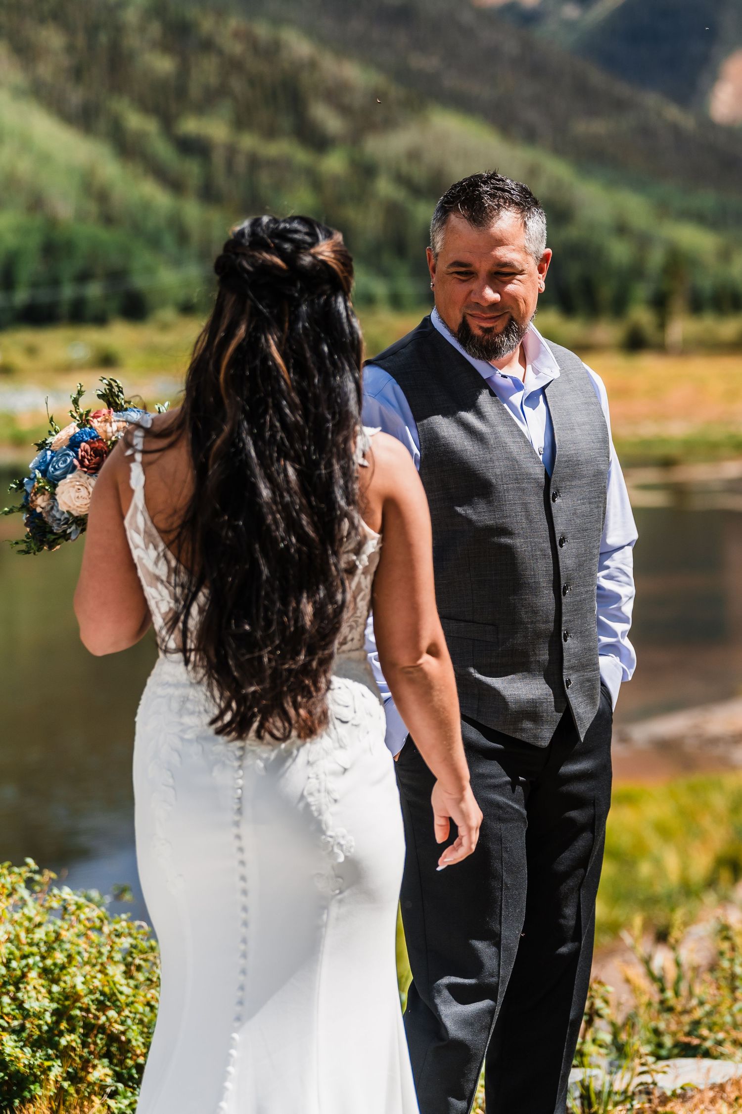 Groom in gray vest smiling at bride in white lace wedding dress holding colorful bouquet by mountain lake.