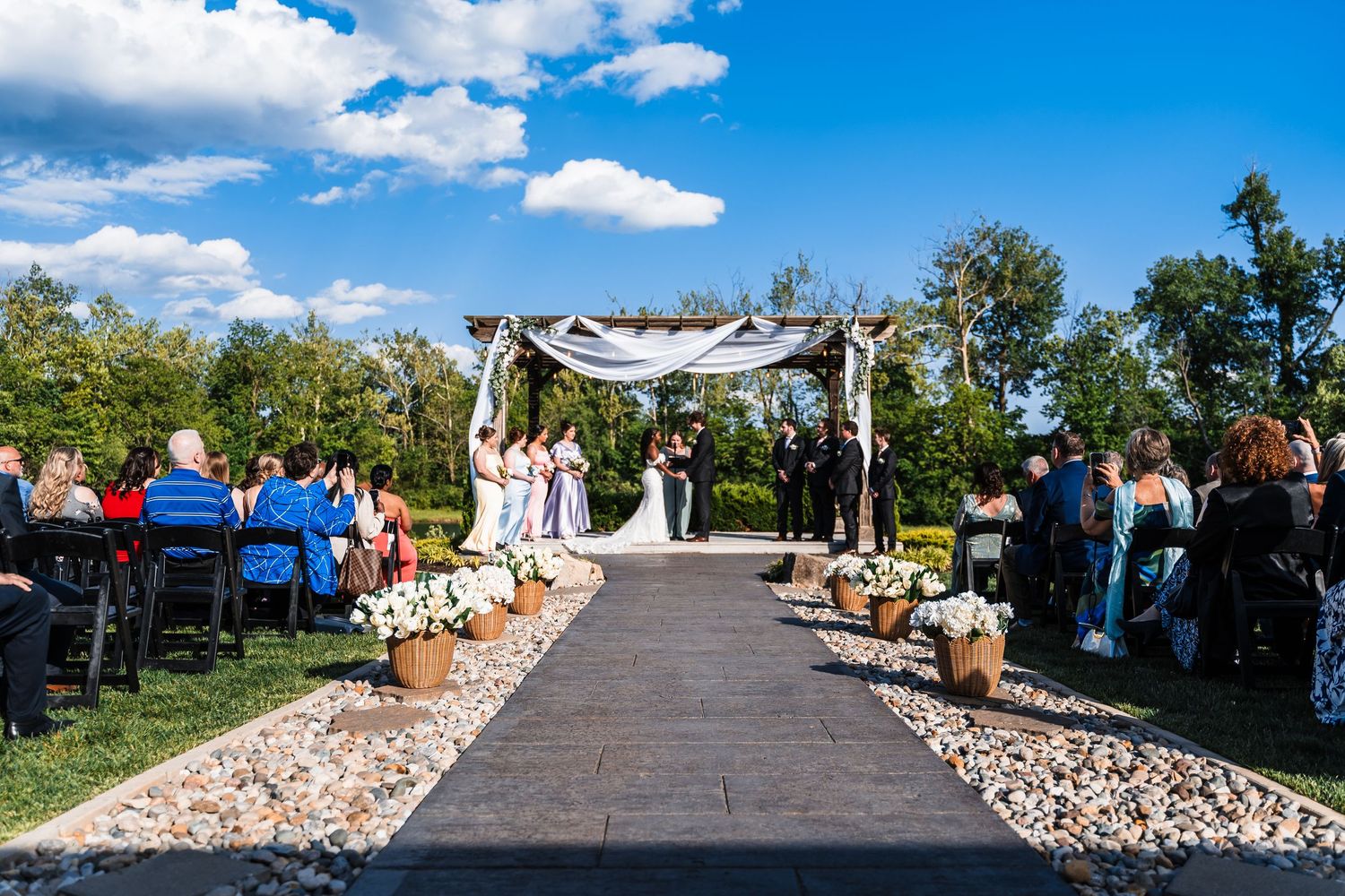 Outdoor wedding ceremony with dark aisle runner, floral arch, and guests seated under blue sky.