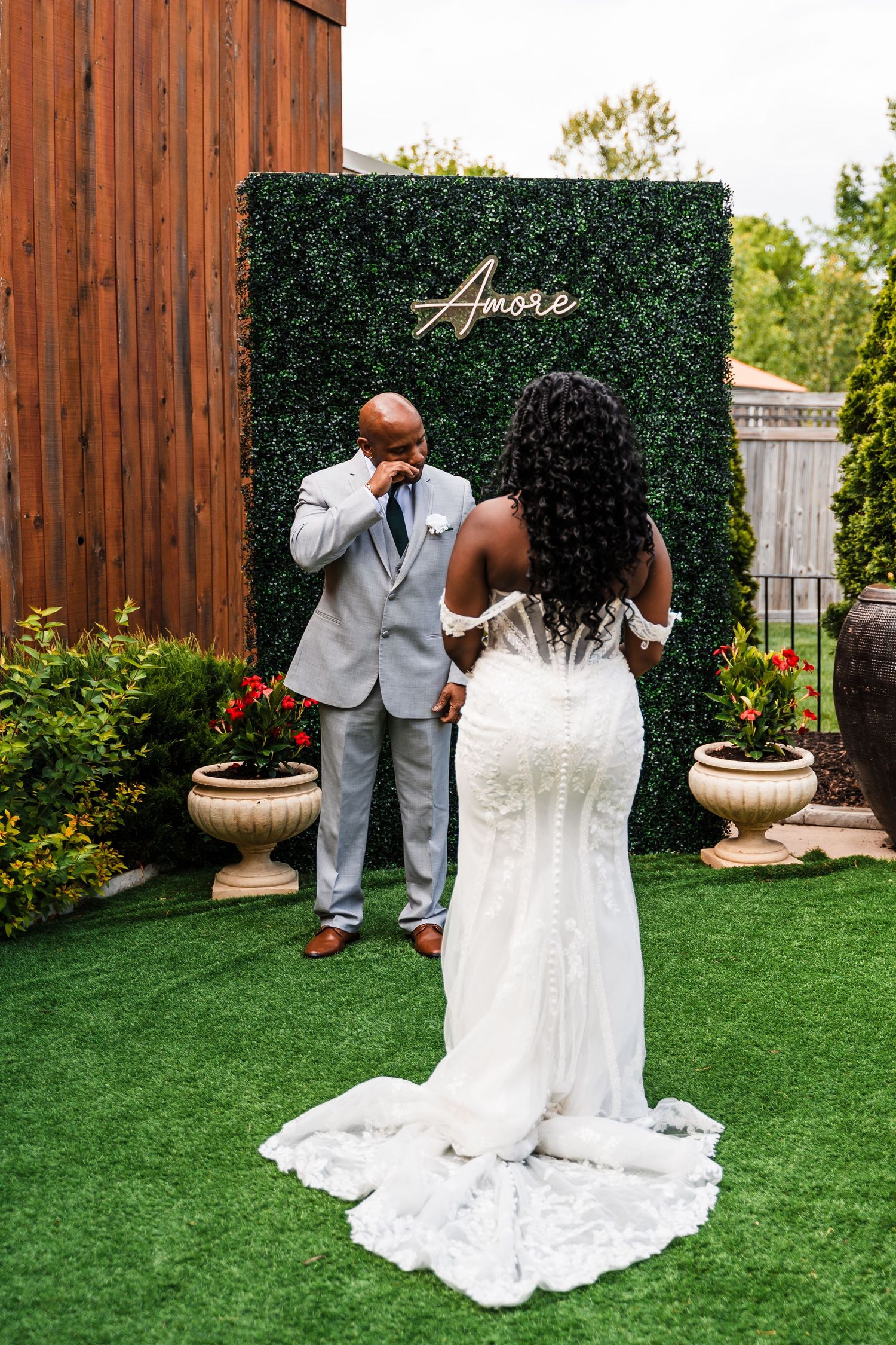 Emotional groom in gray suit wipes tears seeing bride in lace gown during first look at green hedge backdrop.