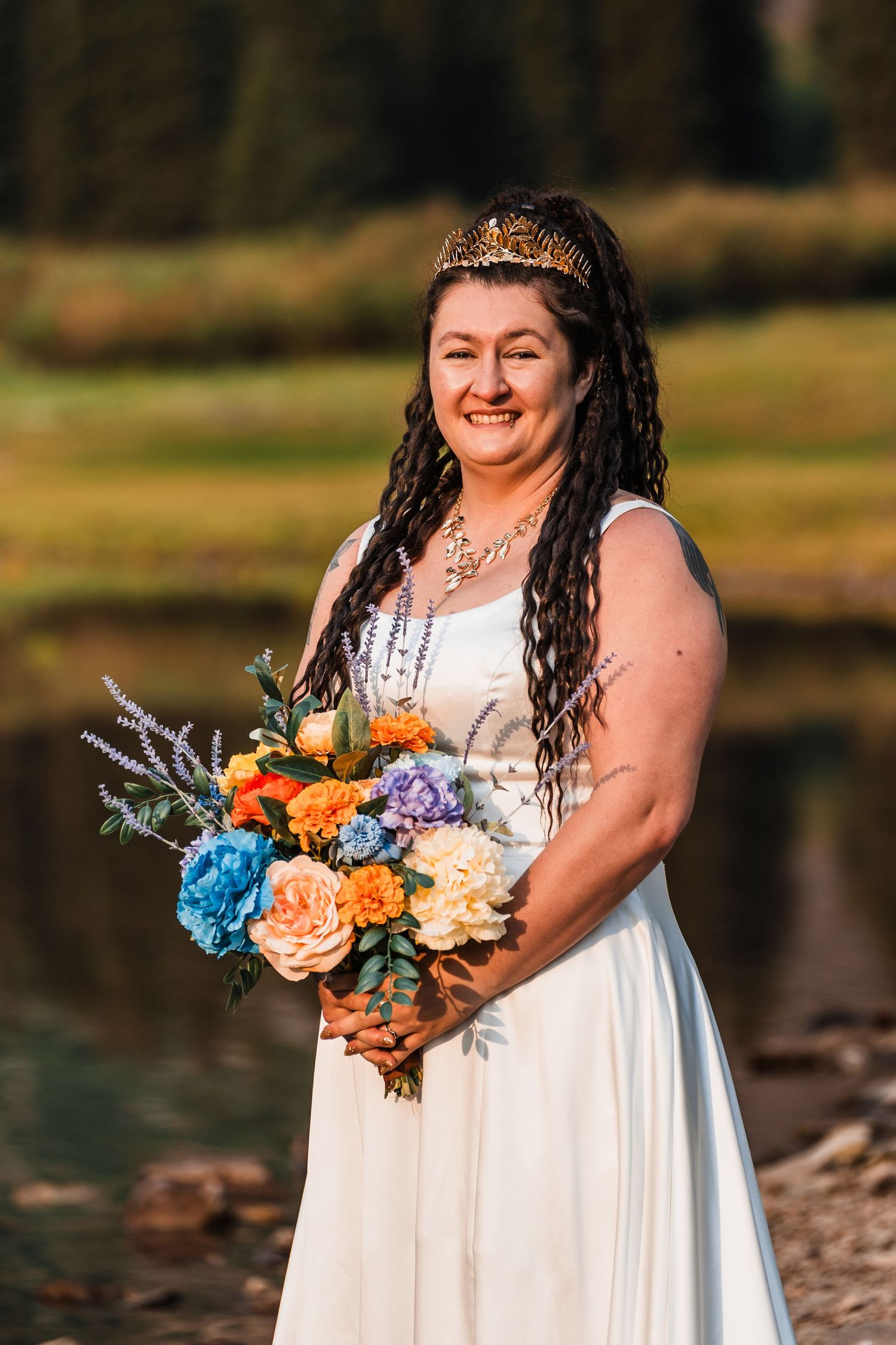 Smiling bride with dreadlocks and gold crown holding vibrant blue, orange, and peach bouquet outdoors.