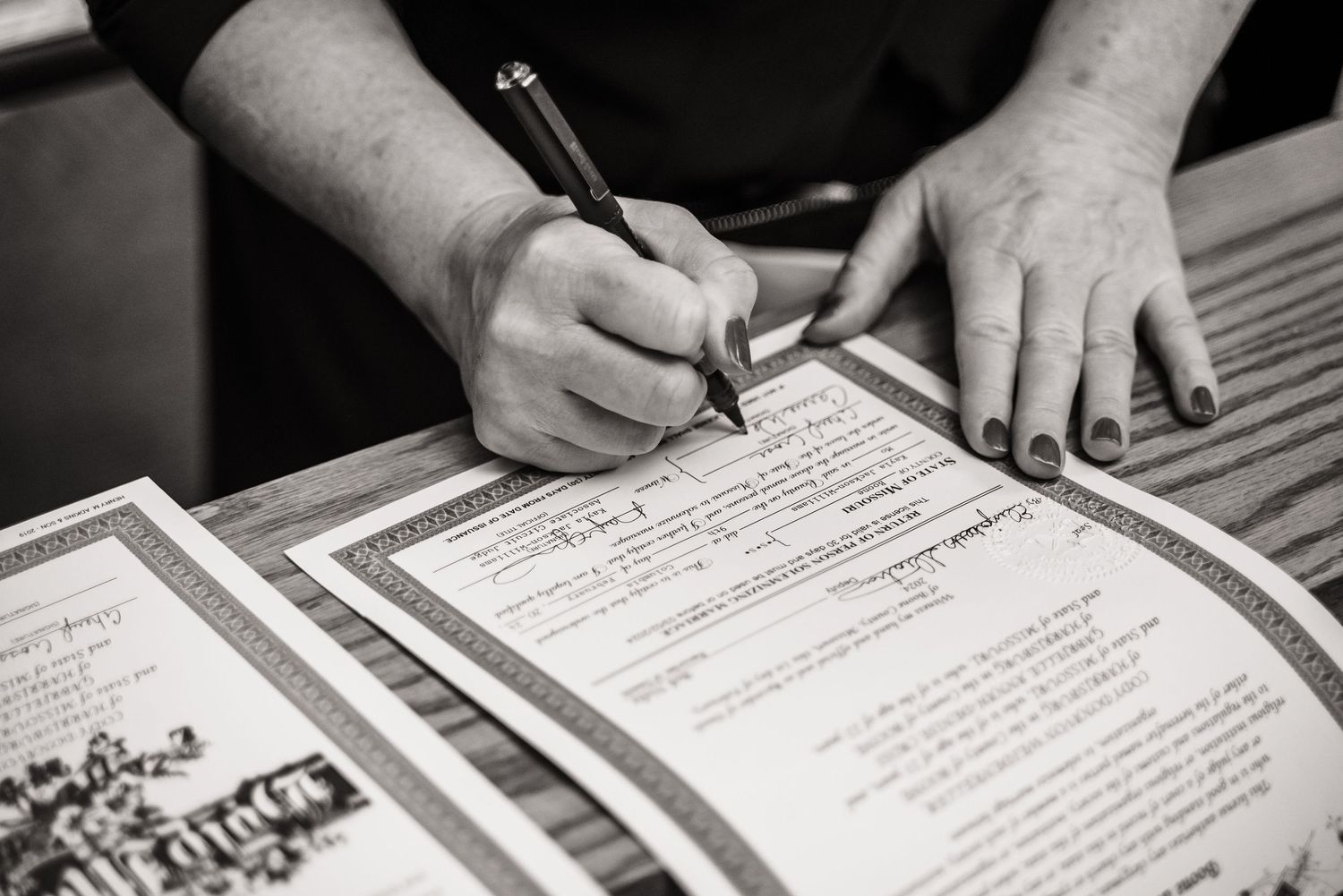 Black and white close-up of hands signing paperwork with a pen on printed documents.